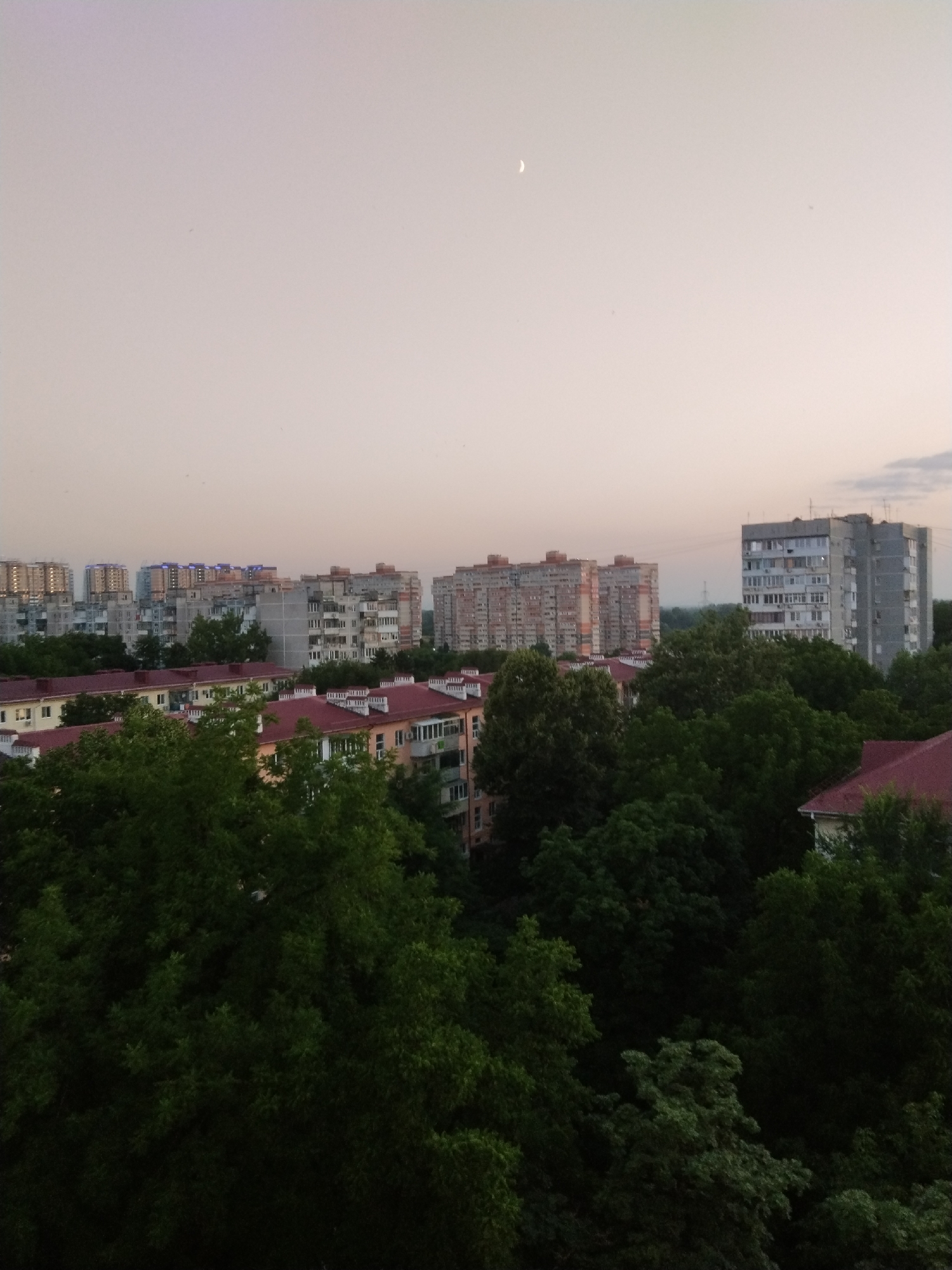 The image depicts a cityscape with a serene sky. In the foreground, lush green trees dominate, obscuring much of the lower portion of the scene. Above the trees, residential buildings of varying heights and architectural styles are visible. Some buildings are light-colored with multiple floors and numerous windows, while others are taller and more modern, with some featuring illuminated details. In the distance, the sky is a soft gradient of pale pink and grey, with a crescent moon faintly visible. The overall impression is of an urban environment at dusk or dawn.