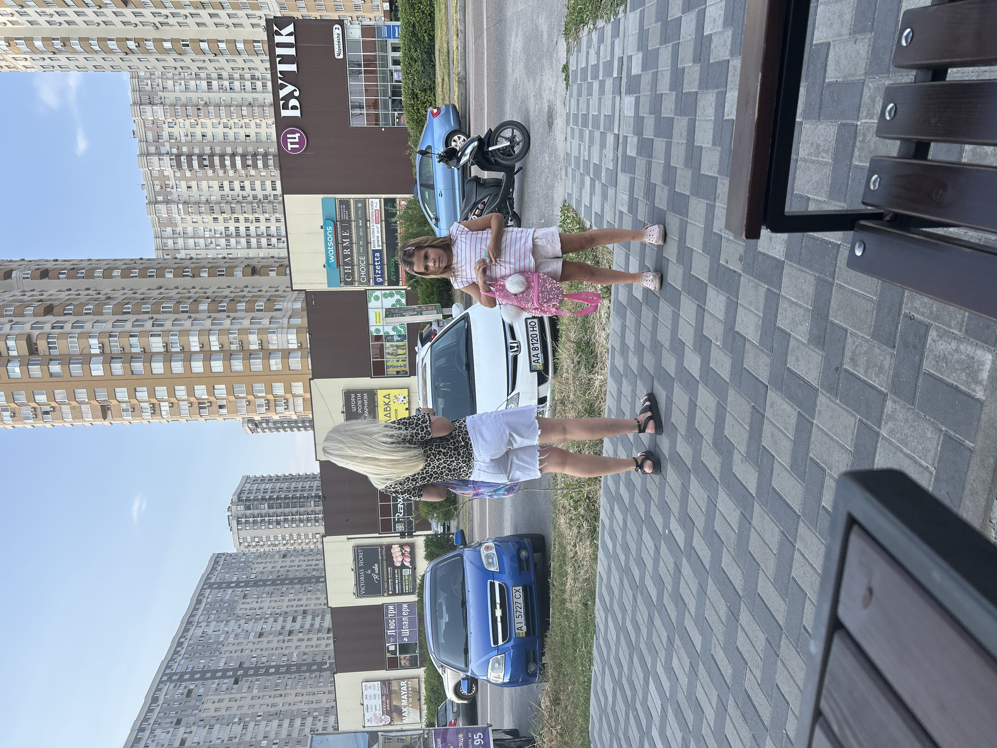 A medium shot captures a woman and a young girl standing on a paved sidewalk in front of a shopping center in Brovary, Ukraine. The woman, with long blonde hair, wears a leopard-print t-shirt and white denim shorts with frayed edges, and black sandals. She is looking down at her phone. The girl, with brown hair, is wearing a striped t-shirt and white shorts, and light pink clogs. She holds a pink backpack with white pom-poms and a phone. Behind them, several cars are parked, including a blue Chevrolet sedan with license plate AI 5727 CX. A black scooter is parked next to a white car. The shopping center has multiple signs for businesses, including "Watsons", "Charme", and "Butik". Tall apartment buildings are visible in the background. The sky is clear with a light haze.