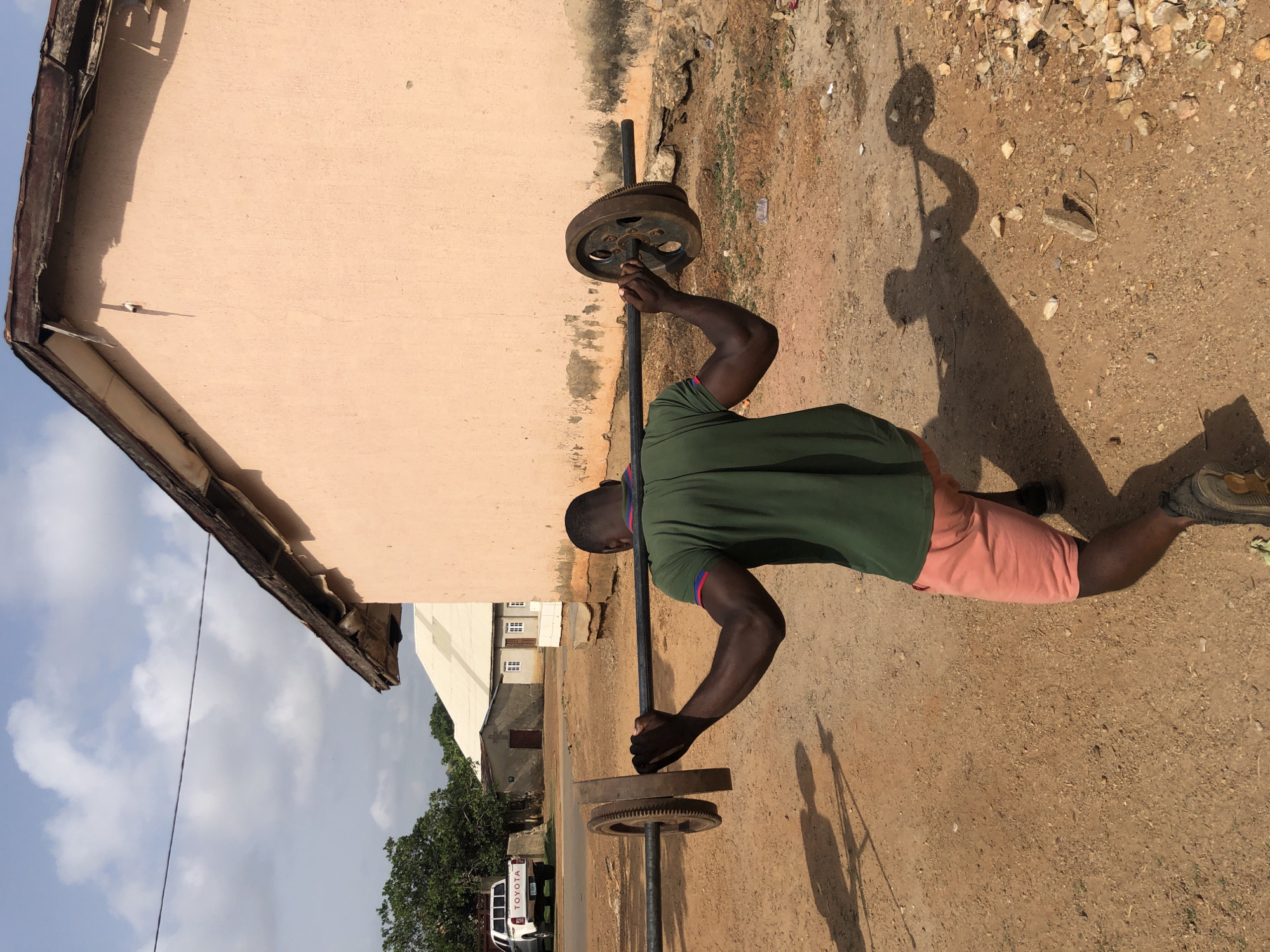 An outdoor scene in Akwanga, Nigeria, captures a man engaged in a physical exercise under bright daylight. The man, with dark skin and an athletic build, is seen from behind, wearing a green t-shirt with red and blue trim on the sleeves, and peach-colored shorts. He is holding a long, black metal bar with both hands, which is weighted on both ends by heavy, dark metal plates, one of which prominently features gear-like teeth. His posture suggests he is in the middle of lifting or lowering the weight, possibly performing a deadlift or a similar strength training exercise.
The setting is characterized by dry, reddish-brown dirt ground scattered with small rocks and gravel. A long, distinct shadow of the man and the weight bar stretches across the ground to his right, indicating strong, direct sunlight, likely morning or late afternoon. To the man's left is a light peach-colored building with a dark, worn roof line. In the background, across a dirt road, are more modest buildings, including a white one with multiple small, rectangular windows, and lush green trees. A white pickup truck is parked in the distance, with the word "TOYOTA" clearly visible on its tailgate. The sky above is partly cloudy.