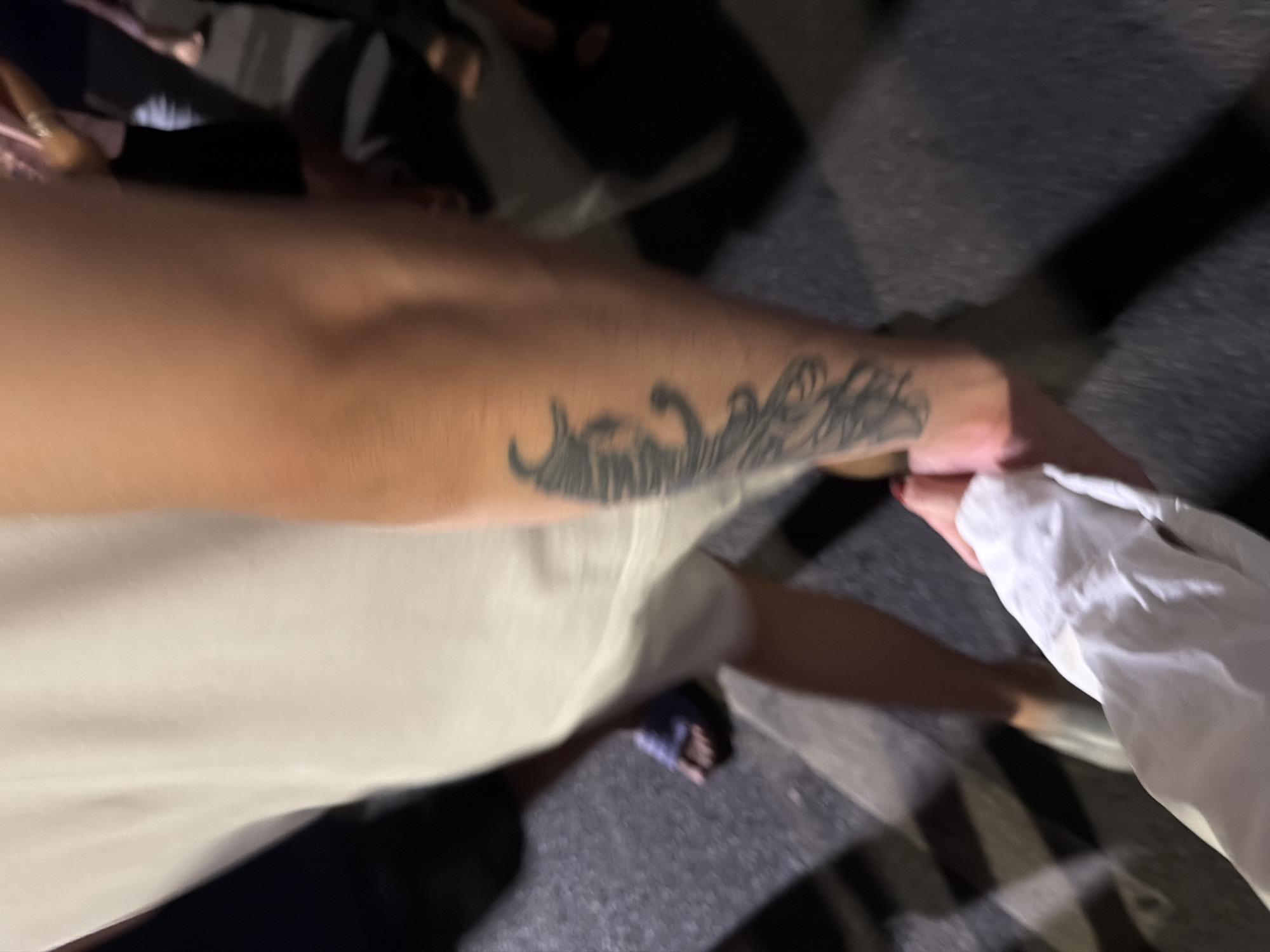 An arm with a tattoo of wavy lines is holding a white fabric. A person wearing a white shirt and blue sandals is visible. The background shows a street with dark pavement. The location is unknown and cannot be determined from the image.