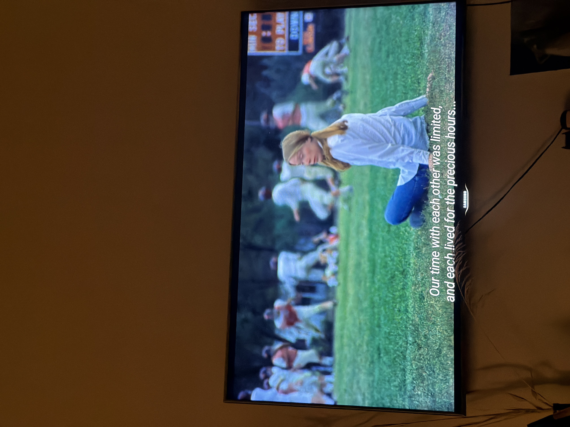 Here's a description of the image:
The image shows a television screen displaying a scene of a movie or show. The scene appears to take place on a grassy field, likely a football field, with several football players in the background, some blurred due to the depth of field. The focus of the scene is a woman kneeling on the grass. She is wearing a white shirt and blue jeans. Text overlay is visible at the bottom of the screen, reading "Our time with each other was limited, and each lived for the precious hours..." The brand of the television, "SAMSUNG," is also displayed. The television is in a room against an off-white or light beige wall. There are black wires visible, connected to the TV and possibly other devices. A portion of what appears to be a framed picture or other wall hanging is visible in the upper right corner. The image is sideways. It is unlikely this image is in Tbilisi, Georgia.