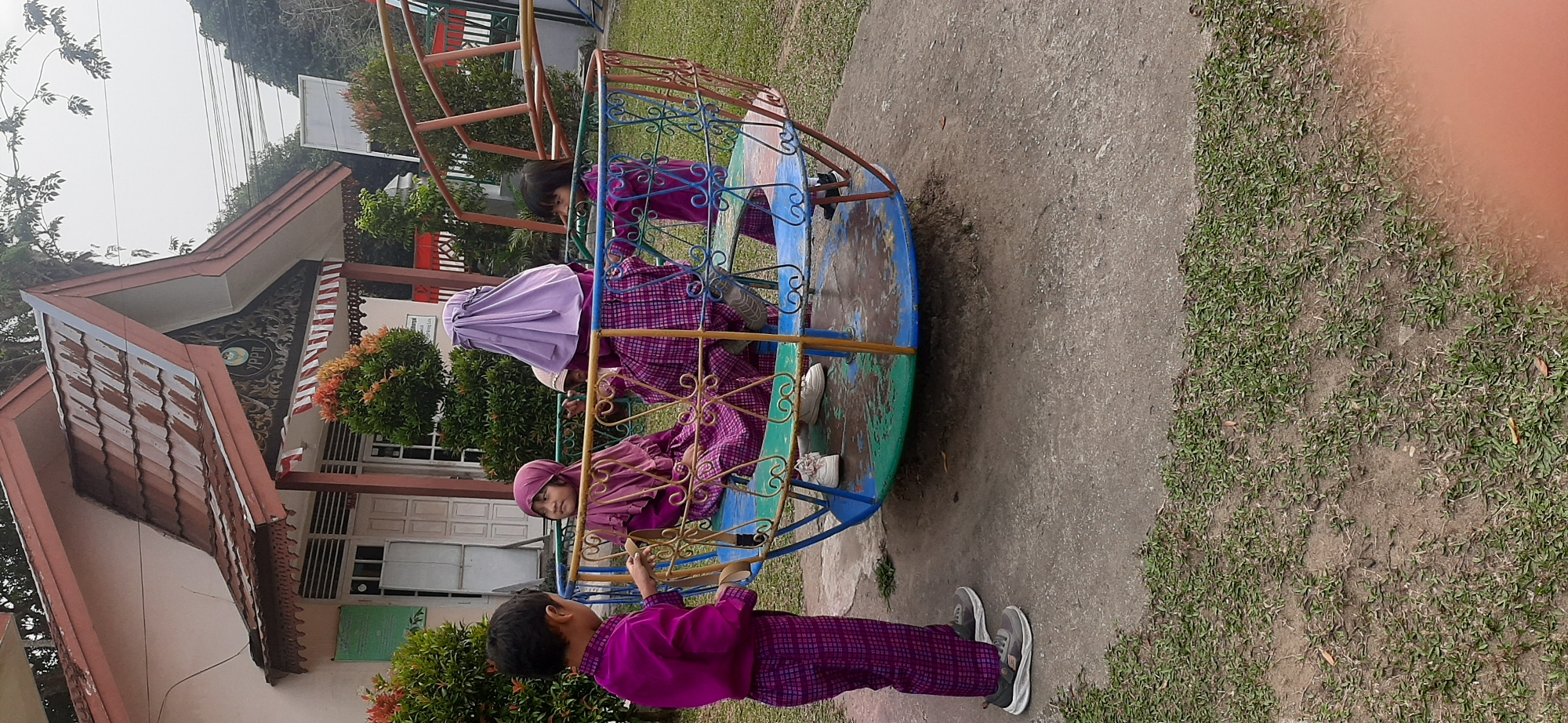 An outdoor scene depicts four children playing on a multi-colored merry-go-round, with a building in the background. The building has a distinctive tiled roof and decorative carvings above the entrance. One child, a boy wearing a purple tunic and checkered pants, stands to the left of the merry-go-round, holding onto one of its poles. Three children are seated on the merry-go-round. Two girls wear purple hijabs and matching patterned tunics and pants, while another child, with dark hair and wearing a purple tunic, is also seated. The merry-go-round itself is a circular metal structure with colorful poles and intricate scrollwork. The ground around the merry-go-round is paved with concrete and surrounded by grass. The overall setting appears to be a schoolyard or playground. The building's architecture and surrounding vegetation suggest a location in a warm climate, possibly Southeast Asia. The presence of the "YPPII" logo on the building indicates it is likely a facility of Yayasan Pendidikan dan Perguruan Islam Indonesia. Based on the architectural style and the signage, the location is very likely the YPPII school in Jambi City, Indonesia.