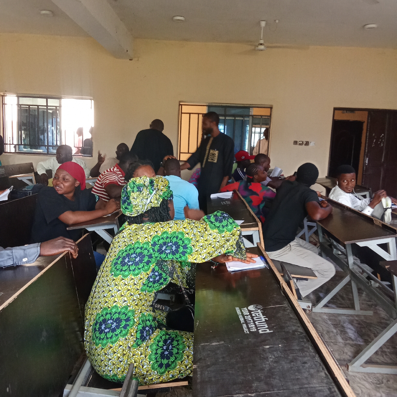The image depicts a bustling daytime scene inside a classroom or training hall located in Akwanga, Nigeria. Numerous individuals, predominantly adults, are seated at wooden desks, suggesting an educational session, workshop, or community meeting.
In the foreground, a woman in a vibrant green, yellow, and blue patterned dress and matching headwrap is seated, facing forward. Another woman to her left, wearing a red headwrap and a black top, is smiling broadly. To the right, a man in a white shirt is seated, looking towards the camera. Throughout the room, other attendees are seated at desks, some appearing attentive, others engaged in conversation. A man in a black tunic with gold embroidery stands near the center-back, possibly an instructor or speaker, addressing the group.
The room is brightly illuminated by natural light entering through windows fitted with metal bars on the left side. The walls are light-colored, and a white ceiling fan is visible overhead. The furniture consists of simple wooden desks and chairs.
Notable details include text visible on a desk in the foreground: "Tetfund" logo, "TETFund", "YEAR 2017/2018/19", and "FUNDED NORMAL PROJECT", indicating the source of funding for the project or facility. Above a door in the background, the word "STORE" is legible. The general mood appears engaged and positive, with several individuals showing smiles or attentive expressions.