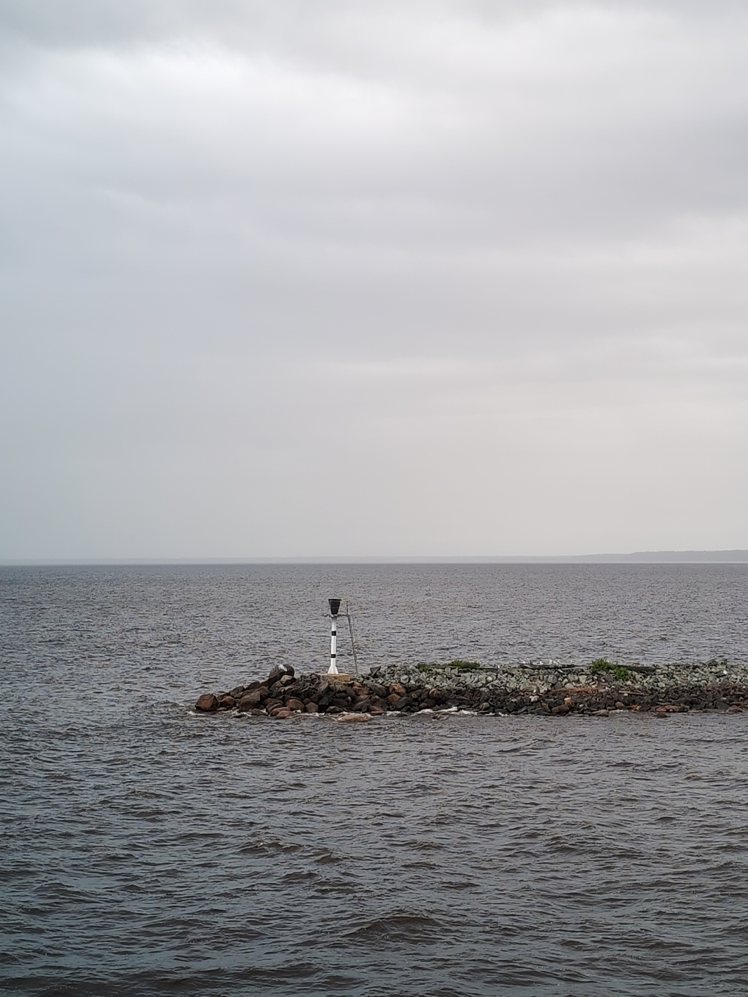 The image shows a body of water under an overcast sky. In the middle of the frame, a rocky breakwater extends into the water, topped with some vegetation. A black and white striped marker stands on the breakwater. The water is dark, and the horizon is visible in the distance. The location is Ozernoe Ust'e, Russia.