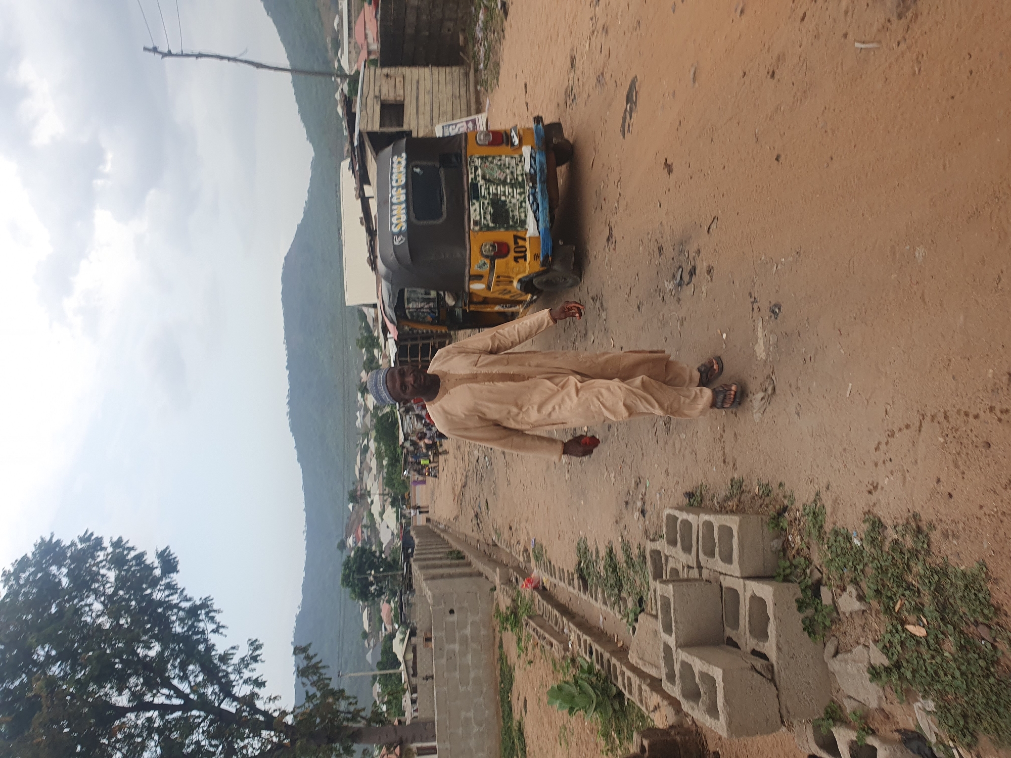 The image depicts an outdoor scene in what appears to be a semi-urban or rural area of Lokoja, Nigeria, under an overcast sky. A man, dressed in a light-colored traditional long garment and a kufi cap, stands on a reddish-brown dirt road. His gaze is directed towards the viewer.
Behind the man, on the dirt road, is a yellow and black three-wheeled auto-rickshaw. The side of the vehicle clearly displays the text "SON OF GRACE", and the number "107" is visible on its rear. To the man's right and extending into the foreground, several grey concrete blocks are stacked on the ground amidst sparse vegetation.
In the background, a settlement with various buildings, some appearing rustic or under construction, stretches towards the base of a large, tree-covered mountain range. Utility poles with wires are also visible against the cloudy sky and mountainous backdrop. A large tree with dark foliage is prominent on the left side of the frame. The time of day appears to be daytime, possibly late morning or afternoon, given the diffused light.