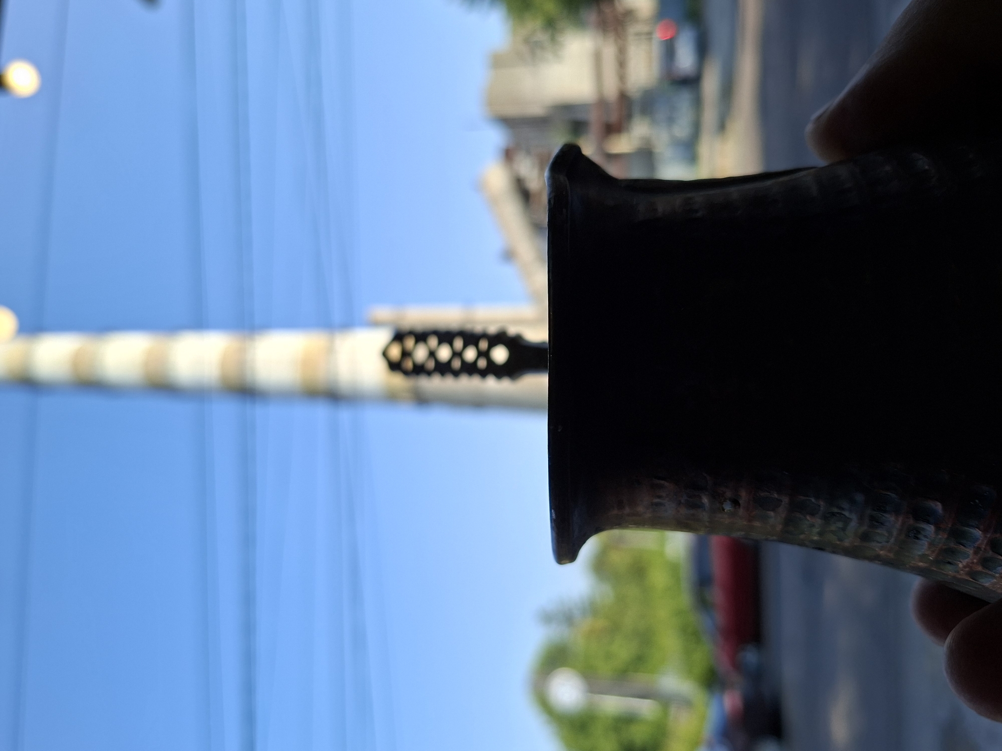 The image shows a close-up view of a dark, textured metal cup or vessel held by a person's hand, with a blurred background. In the background, a tall, striped industrial chimney, likely a power plant or factory stack, is visible against a bright blue sky with thin horizontal power lines. Further in the distance, a blurred cityscape with buildings and trees is visible, along with a red car on a street. The overall scene suggests an urban environment with industrial elements. The specific location within Chișinău, Moldova cannot be determined from the visual information provided.