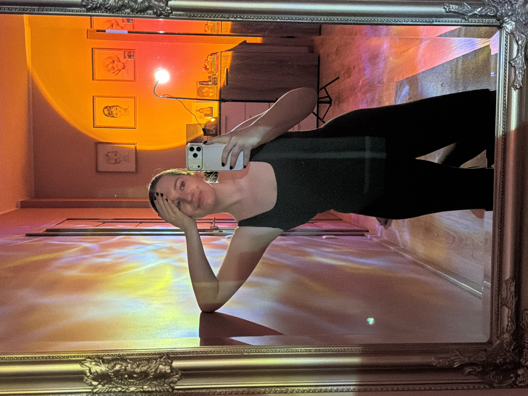 A person is reflected in an ornate, vintage-style mirror. The individual is positioned at an angle, holding a smartphone and appearing to take a selfie. They are wearing black activewear. The background, visible through the mirror's reflection, includes a room with artwork on the walls, a floor lit with yellow and orange light patterns, and a cabinet or storage unit. The scene seems to be indoors. There is no specific information in the image confirming the location in Dreiliņi, Latvia, or any indication of activity in that city.