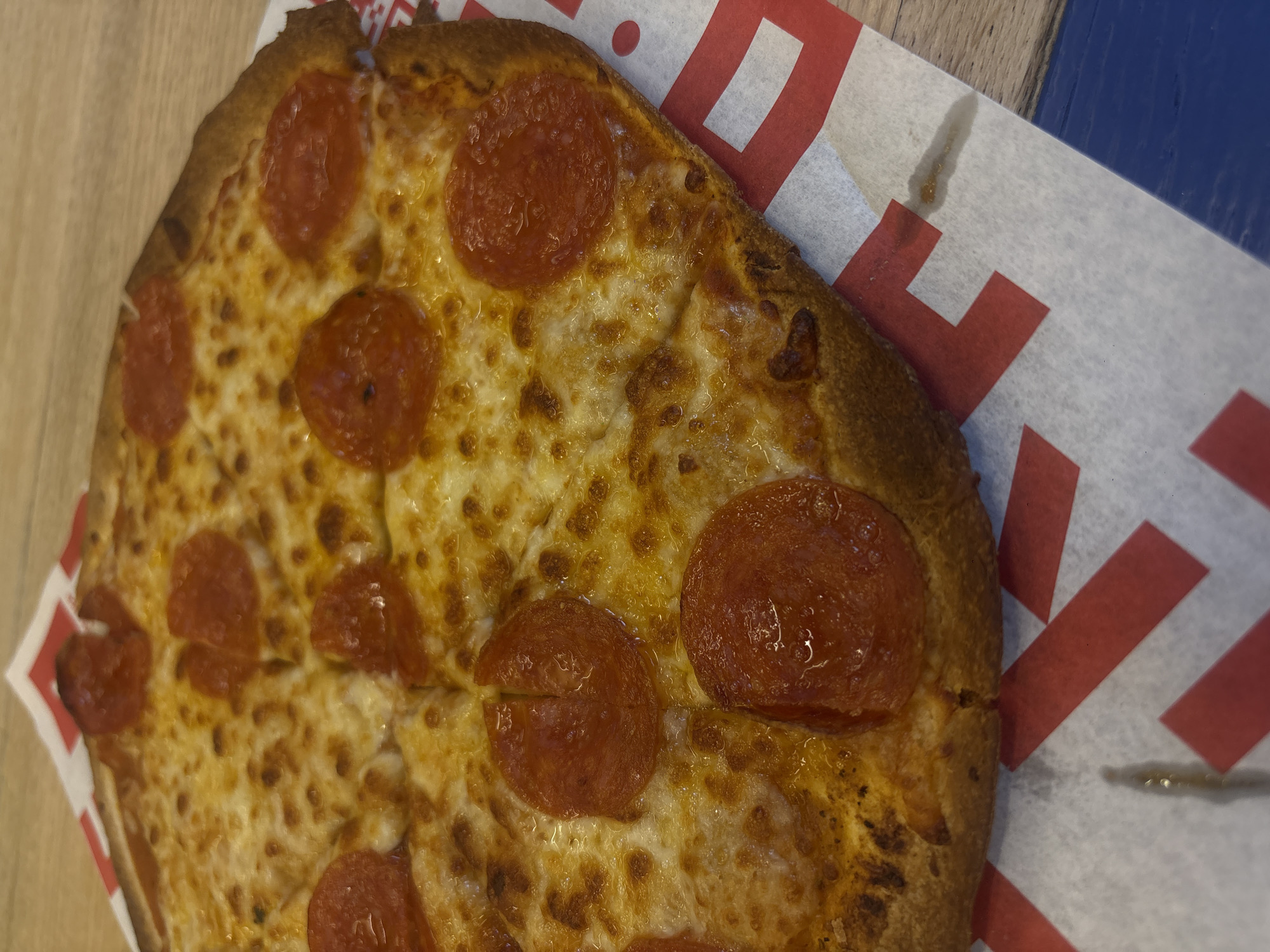 A top-down, close-up shot shows a slice of pepperoni pizza on a paper plate with red and white geometric shapes. The pizza has a golden-brown crust and melted cheese with several pepperoni slices. The paper plate is on top of a wooden surface that appears to be part of a table. The location is not identifiable as Mexico City based on the image.