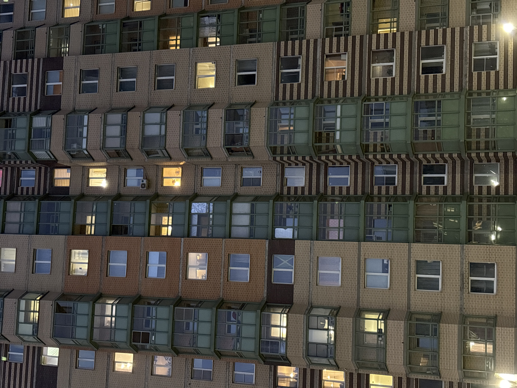 A medium, orthogonal, eye-level shot depicts a multi-story residential building, characteristic of urban architecture in Kudrovo, Russia. The facade is a repetitive pattern of windows and brick panels. Alternating rows have cantilevered balconies or window extensions with green framing. The bricks vary in color, ranging from light tan to reddish brown, with some sections featuring vertical striped patterns in darker tones. Some windows are illuminated from within, suggesting occupants are present. The lighting is ambient and appears to be either dusk or night. No people are visible in the image.
