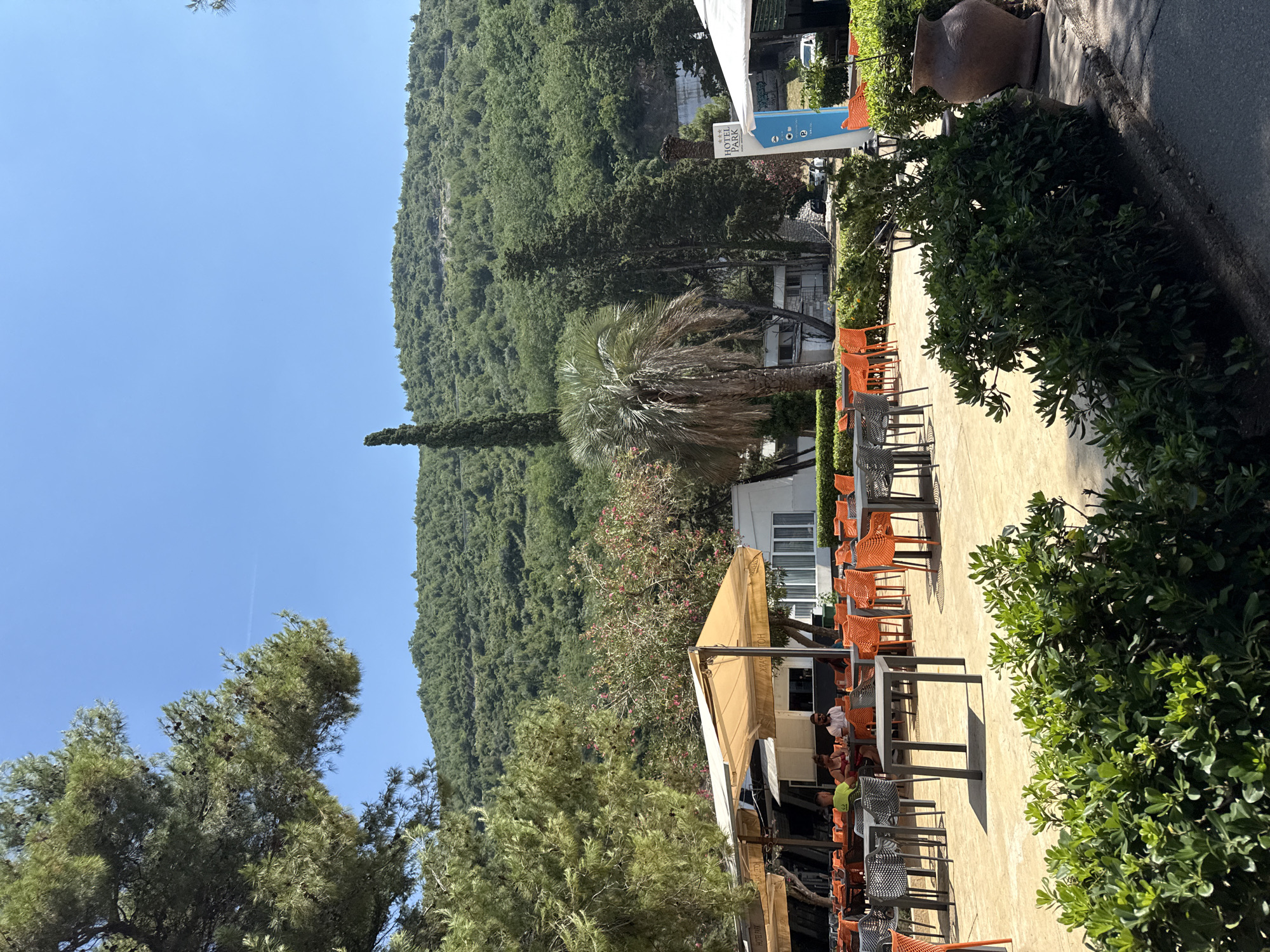 A bright, sunlit outdoor seating area of a restaurant is depicted, likely part of the Hotel Park in Budva, Montenegro. The scene faces a lush, green, mountainous backdrop under a clear blue sky. In the foreground, meticulously maintained green bushes line a paved walkway. Beyond the bushes, a series of tables and chairs are arranged on a light-colored paved surface. Many of the chairs are a vibrant orange, while others are dark grey. Some tables are occupied by patrons, including a group of three people seated at one table near the left side of the frame, seemingly engaged in conversation. A large, light-colored umbrella provides shade over a section of the seating area. In the mid-ground, a palm tree stands tall next to a tall, slender cypress tree. A sign for "HOTEL PARK" is visible on the right side of the image, with a blue directional indicator below it. Several white buildings, possibly part of the hotel, are situated behind the seating area. A large, rustic clay pot is visible on the right edge of the frame. The overall atmosphere is relaxed and summery, suggesting a pleasant daytime setting for dining or socializing.