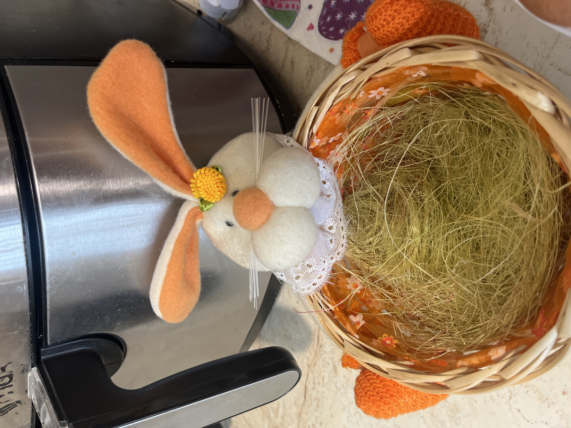 A close-up shows an Easter-themed arrangement on a light-colored countertop. To the left, resting on a brushed metal surface, is a plush rabbit head with orange ears and a yellow flower detail. To the right, a wicker basket is filled with light-colored straw. The basket has an orange fabric lining with a floral pattern. The bottom of the basket shows a knitted orange detail, possibly carrots. Based on the image, it is impossible to verify the location within the city of Vladaya, Bulgaria.