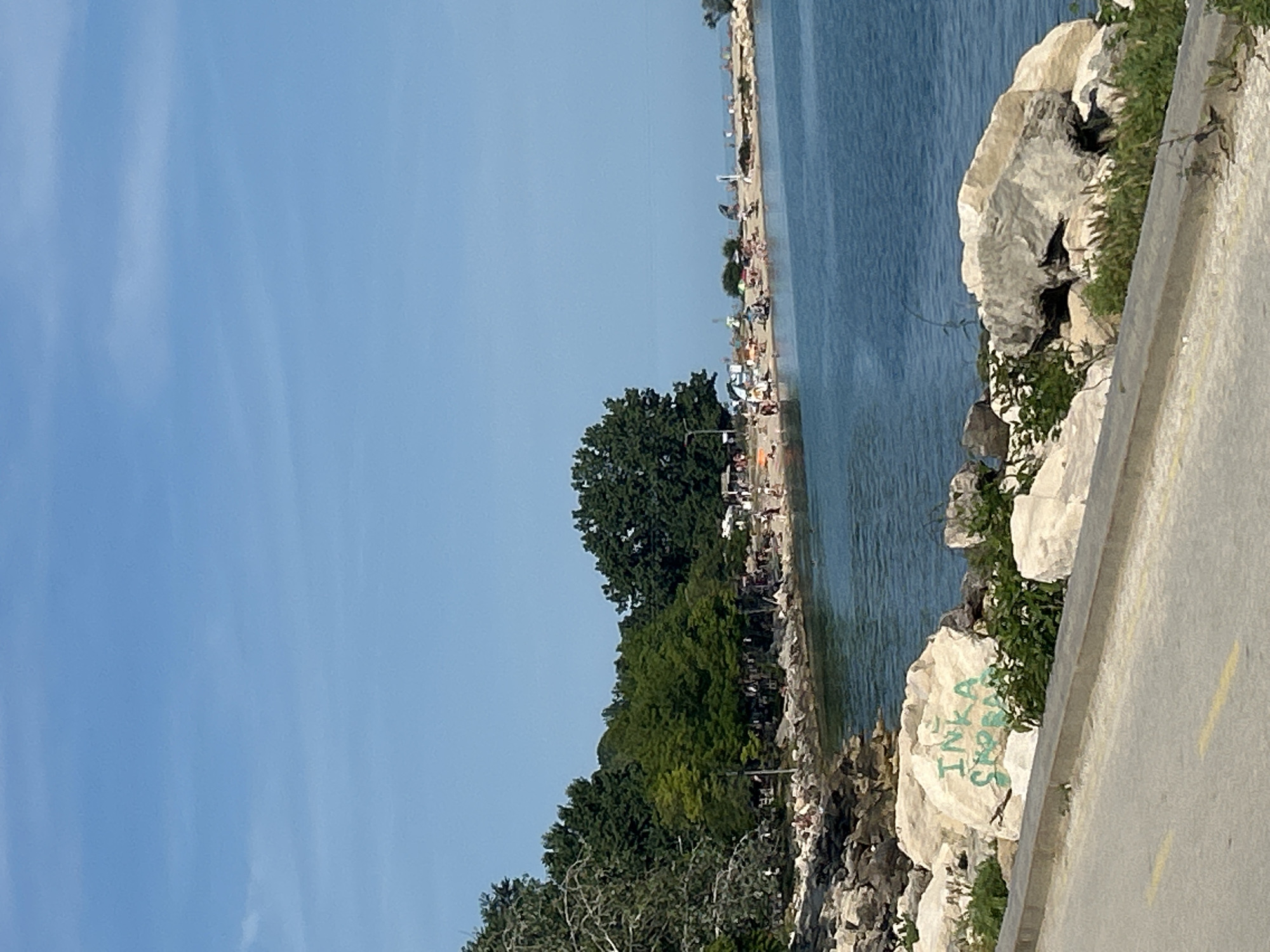 This image depicts a sunlit coastal scene in the city of Unknown, Unknown. In the foreground, a gray asphalt road featuring a partial yellow dashed line runs parallel to a concrete curb. Adjacent to the curb, several light-colored boulders are visible, interspersed with green vegetation. One prominent boulder displays green graffiti in uppercase letters, reading 'INKA' above 'SHOPBAO'. Beyond these rocks, a body of calm, dark blue-green water forms a cove.
The midground showcases a rocky shoreline that transitions into a sandy beach. Dozens of individuals are present on the beach and near the water, many engaged in sunbathing or relaxing under various beach umbrellas. Several small, light-colored structures are scattered along the back edge of the beach. Dense green deciduous trees and bushes form a backdrop, with some taller, darker green trees visible further inland.
The background is composed of a clear, light blue sky with only faint, wispy white cloud formations visible in the upper left. The horizon line where the water meets the sky is discernible on the far right.