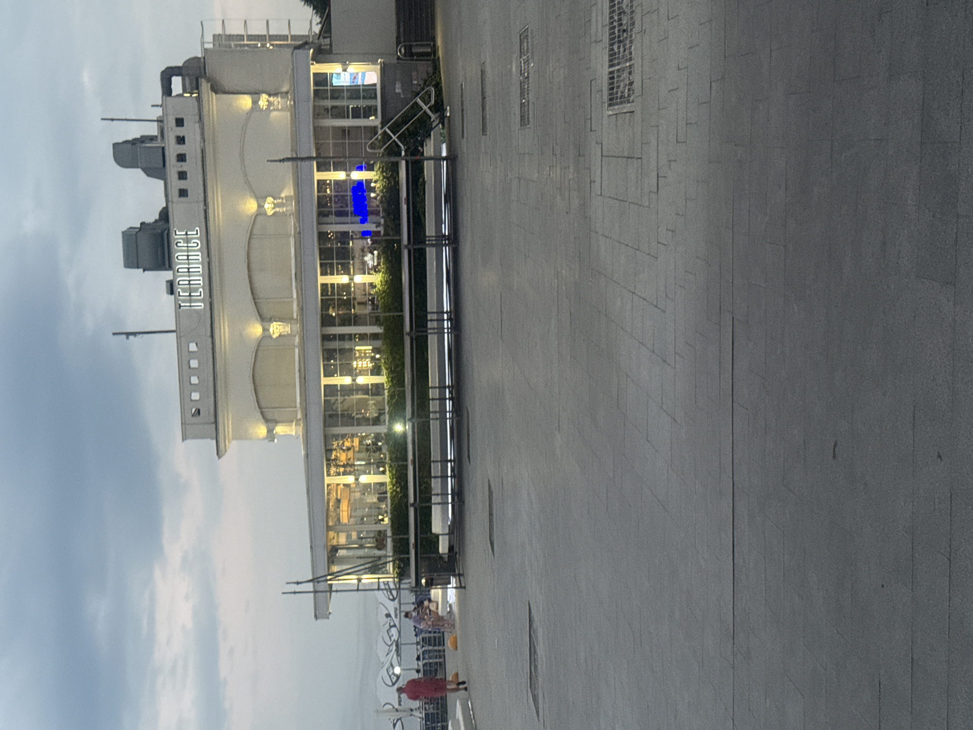 The image shows a building with the word "TERRACE" illuminated in white letters on its facade. The building is a light color and has a modern architectural style with large windows. In front of the building, there is a paved area with several metal tables and chairs. To the left of the building, a person wearing a red shirt and dark pants is walking away from the camera. Further back, other people can be seen near umbrellas. The sky above is overcast and the lighting suggests it is either early morning or late evening. The scene appears to be a public space, possibly a promenade or a waterfront area in Odesa, Ukraine, given the context. The overall impression is of a serene, urban environment at dusk or dawn.