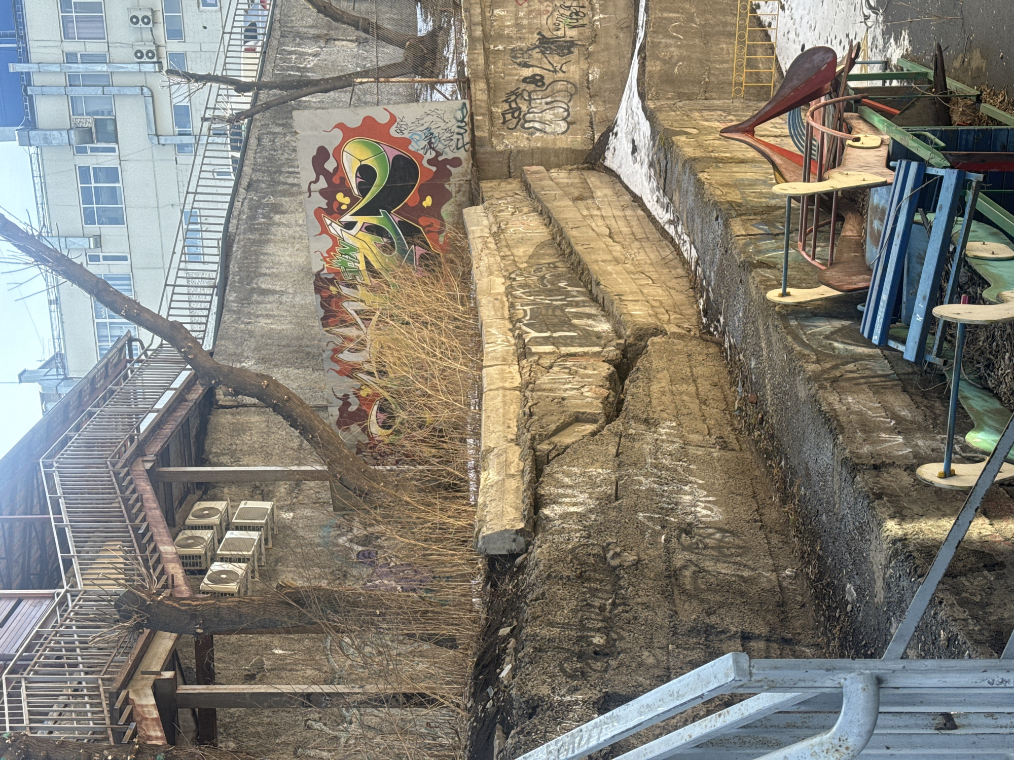 This scene depicts an urban environment, likely in disrepair. A tall building with multiple windows and air conditioning units is visible on the left side of the image, next to a structure covered in graffiti, including what appears to be a stylized number "2." Bare tree branches partially obscure the view of the graffiti. To the right of the building, there's a crumbling concrete structure, possibly a wall or bridge abutment, with significant cracks and weathering. On top of this structure, there are remnants of what appear to be discarded or abandoned playground equipment. The overall impression is one of neglect and decay. Given the prompt's location, this scene is possibly located in or around the city of Krasnoyarsk, Russia.