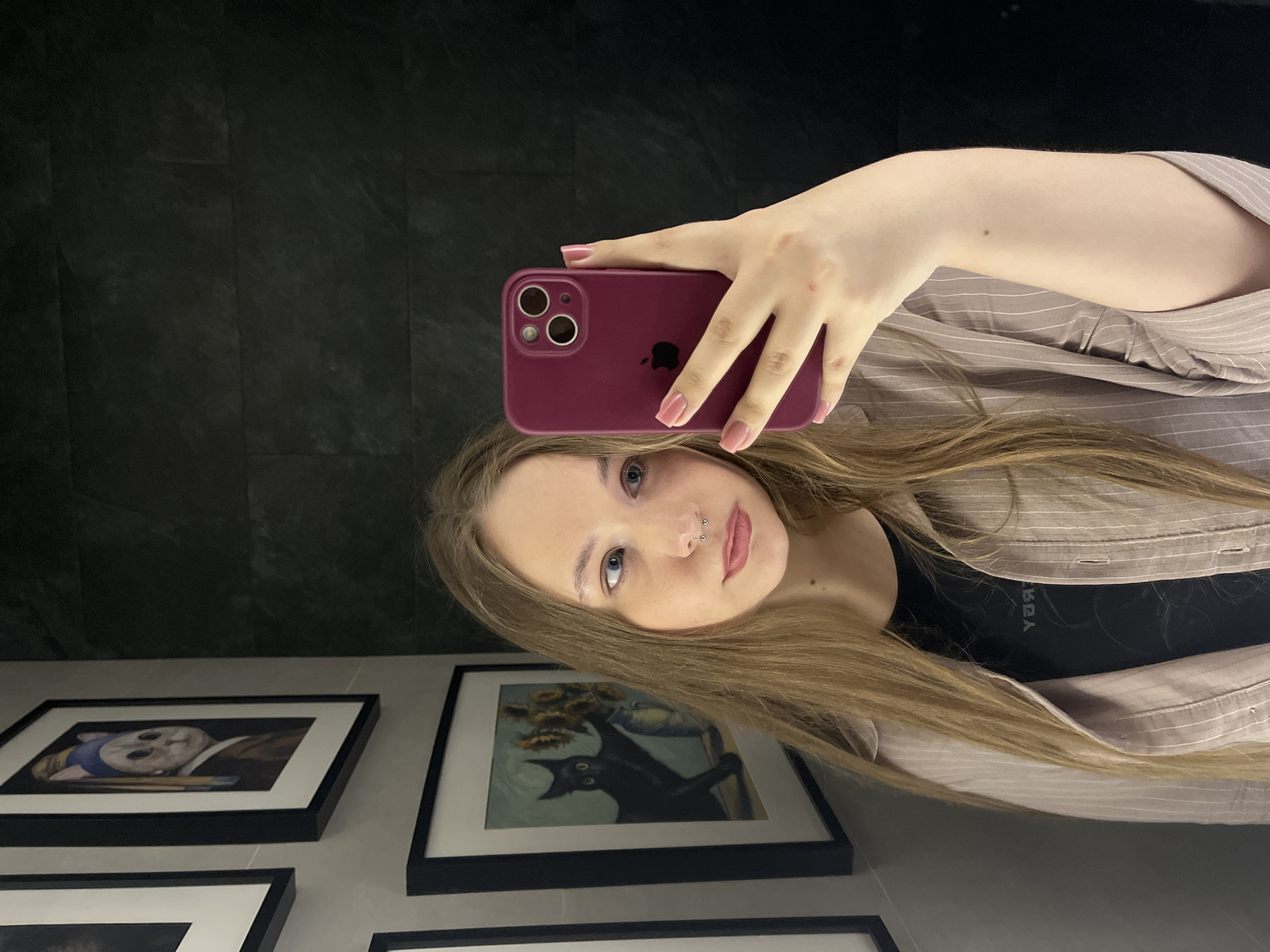 A young woman with fair skin and long, light brown hair takes a selfie with a purple iPhone. She has blue eyes and a nose piercing. She's wearing a beige pinstripe shirt over a black top. The background is a black wall, and there are framed pictures hanging on the wall behind her. The location is Unknown, Unknown.