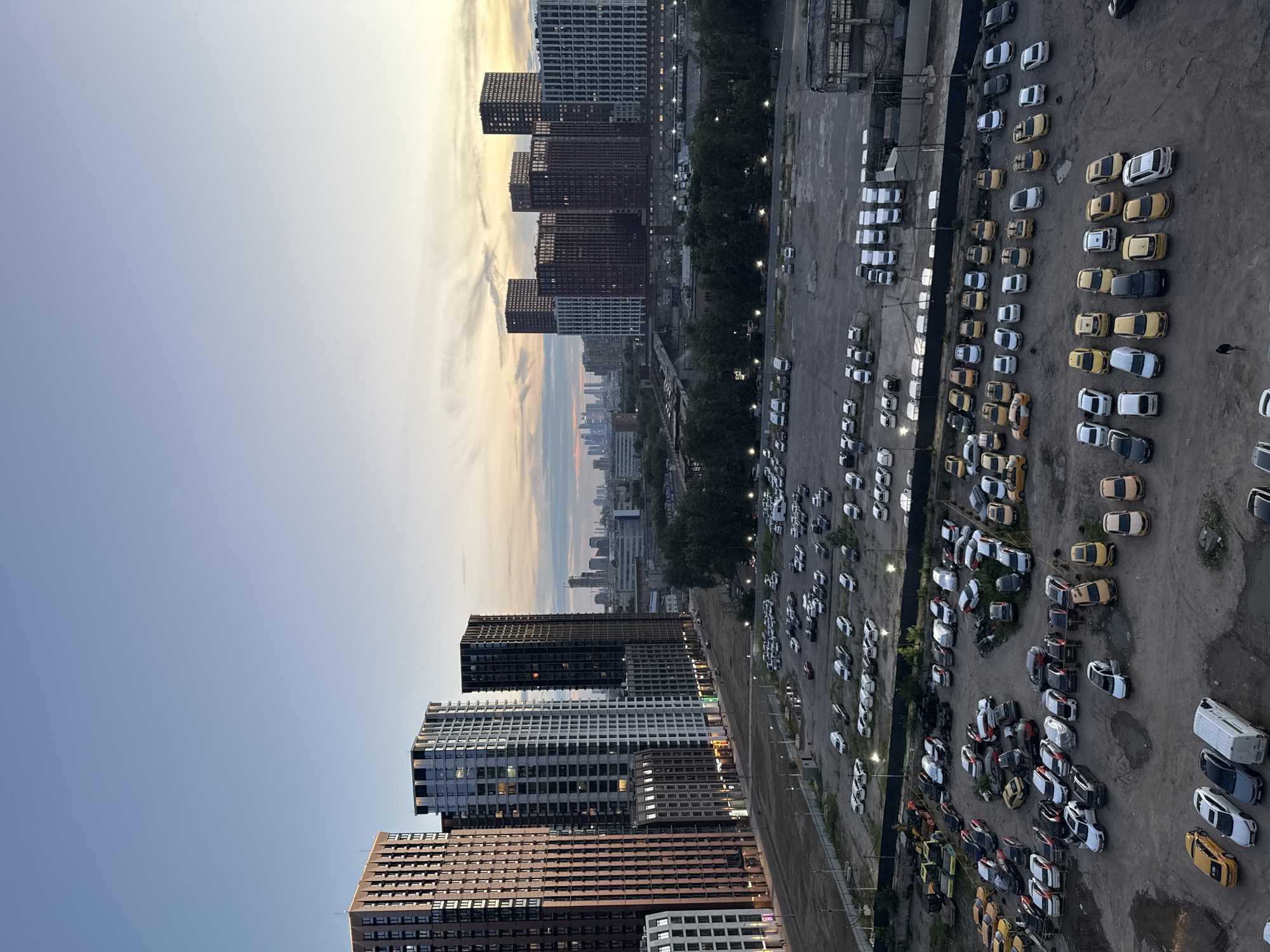 The image depicts an urban landscape at dusk, characterized by a wide sky transitioning from pale blue to hues of orange and yellow. In the foreground, a large, open, unpaved area serves as a parking lot filled with numerous vehicles. These vehicles are predominantly cars, with a variety of colors and models, including what appear to be taxis. There are also a few vans and a construction vehicle visible. The parking lot is bordered by a dark fence. Beyond the parking lot, a row of trees separates it from a complex of modern, multi-story residential buildings. Further in the distance, more skyscrapers and urban structures are visible, forming a city skyline under the twilight sky. The overall impression is a developing urban area with a mix of residential and commercial structures. The location is identified as Bobrovo, Russia. There is one person visible walking in the parking lot.