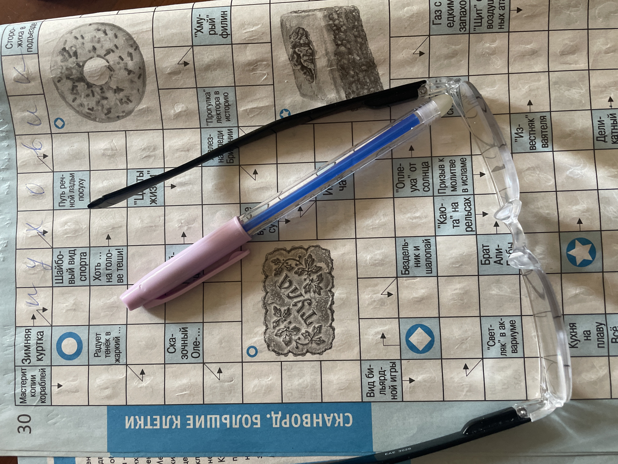 The image shows a close-up of a partially completed crossword puzzle in a newspaper. The crossword grid is visible, with several words filled in and others represented by clues. A pink pen and a pair of clear-framed glasses are resting on top of the newspaper, partially obscuring the puzzle. The newspaper appears to be a local publication, as suggested by the text in Russian. The context of Minsk, Belarus is not derivable from the image. The image captures a mundane scene of someone engaging with a crossword puzzle.