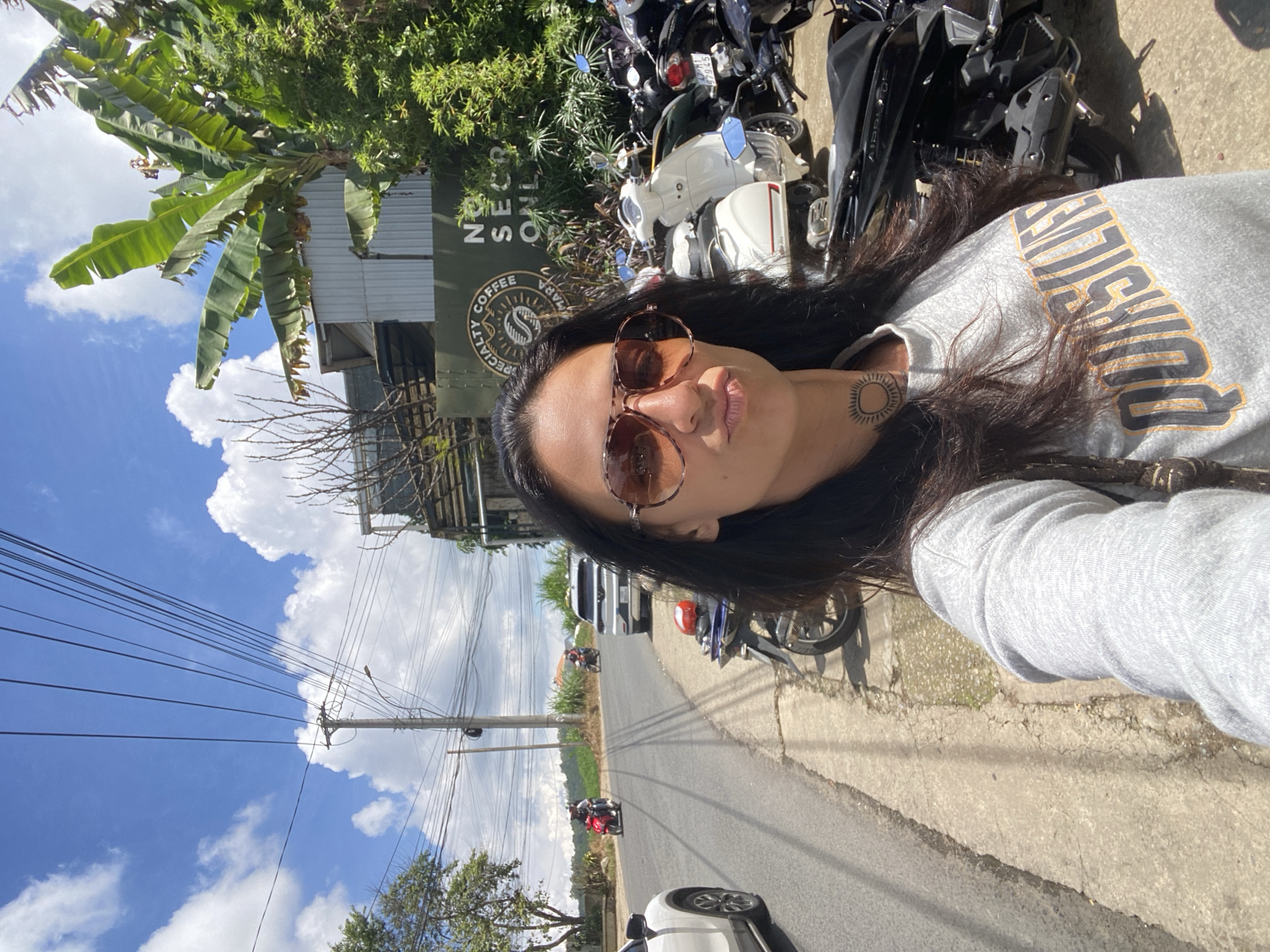 A woman is taking a selfie in front of a specialty coffee shop on a sunny day in Da Lat, Vietnam. She has long, dark hair and is wearing sunglasses and a grey sweatshirt with an orange and black graphic. Behind her, a sign for "NO SECRETS COFFEE" is visible, along with a row of parked scooters and motorbikes. The street is paved, and there is a sidewalk on the right. In the background, there are trees, power lines, and a white car. The sky is blue with white clouds.