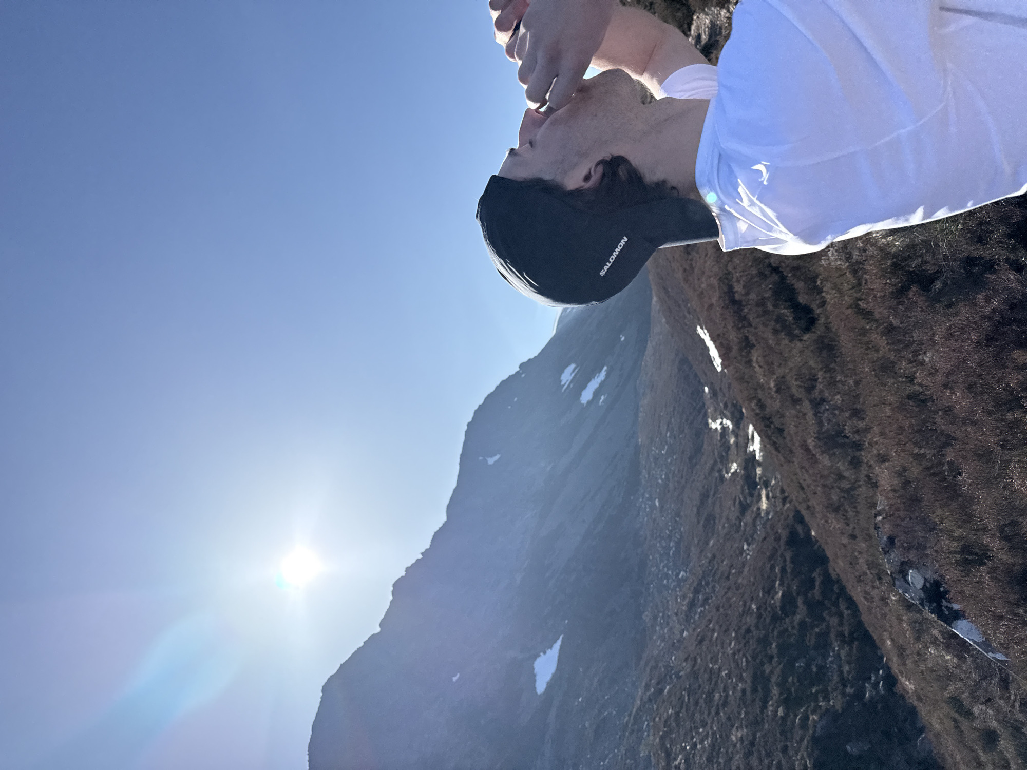 A man wearing a white t-shirt and a black Salomon cap is drinking from his hands. The background consists of a mountain partially covered in brown vegetation with patches of snow. The sky is clear and blue, and the sun is shining brightly. The location is Unknown, Unknown.