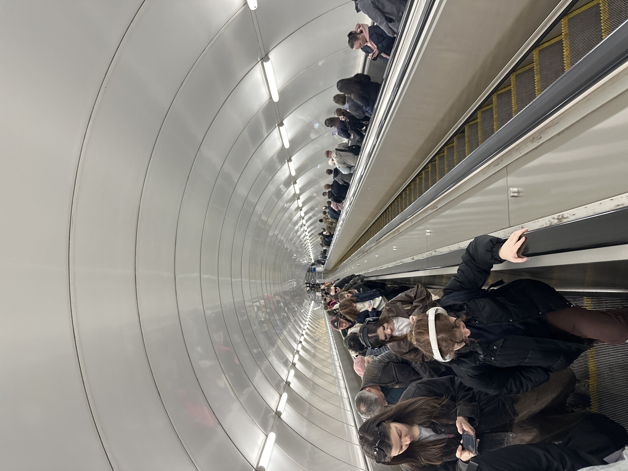 The image captures a long, deep escalator tunnel, characteristic of a metro station in Saint Petersburg, Russia. The perspective is tilted, showing the escalators running almost vertically from the foreground into the distance. The tunnel walls are uniformly light gray, curved, and segmented by subtle vertical lines, creating an architectural rhythm. Numerous horizontal fluorescent tube lights illuminate the tunnel, spaced regularly along the ceiling.
Multiple people are visible on both the left and right escalators, extending far into the background. They appear to be primarily adults, dressed in various winter or cool-weather garments such as jackets, coats, and hoodies. Many individuals are standing on the escalator steps, holding onto the dark handrails. Some are looking forward, others are engaged with their smartphones. In the immediate foreground, a person with long dark hair holds a phone, and another person wears prominent white over-ear headphones. The escalator steps feature alternating dark gray and bright yellow sections.
The scene depicts the daily activity of commuters or travelers moving through a busy underground transport system. The deep tunnel and the style of the escalator are consistent with the known characteristics of the Saint Petersburg Metro. The lighting is artificial and uniform, suggesting an indoor environment where the time of day is not apparent. No discernible text is visible in the image.