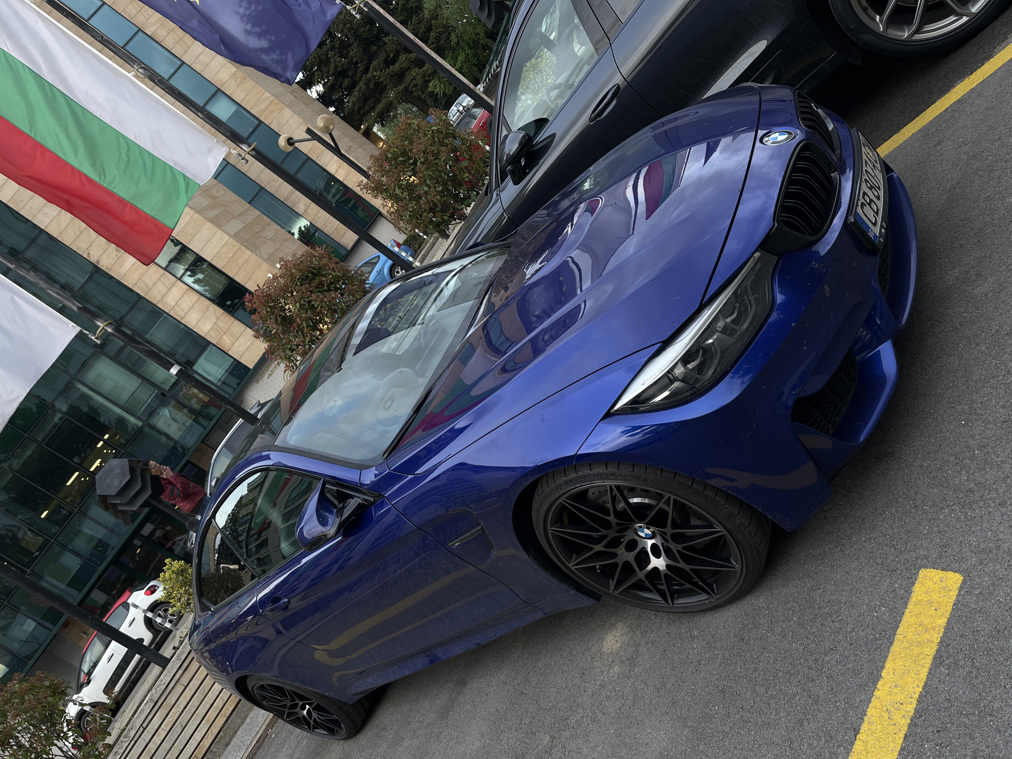 A vibrant blue BMW M4 coupe (F82 generation) is prominently parked diagonally across an outdoor asphalt lot, serving as the central subject of the image. The car showcases its aggressive black grille, sleek headlights, and black multi-spoke alloy wheels with "MICHELIN" visible on the tires. Its reflective surface captures the surrounding environment, including a clear reflection of a white, green, and red striped flag (the flag of Bulgaria) and a blue flag adorned with yellow stars (the European Union flag).
Adjacent to the BMW, a dark-colored SUV is partially visible. In the background, a contemporary building with large glass windows and a light-colored facade rises, reflecting parts of the sky and surroundings. Lush green trees and smaller bushes are interspersed around the building. A person holding a black umbrella is discernible in the mid-ground, suggesting either recent rainfall or protection from the sun. The ground is a dark asphalt lot marked with a bright yellow parking line. The BMW's license plate, displaying "CB 8074 AH", provides a clear location cue, confirming the setting as Sofia, Bulgaria, which aligns with the national and EU flags. The scene depicts an urban, commercial, or administrative area during daytime.