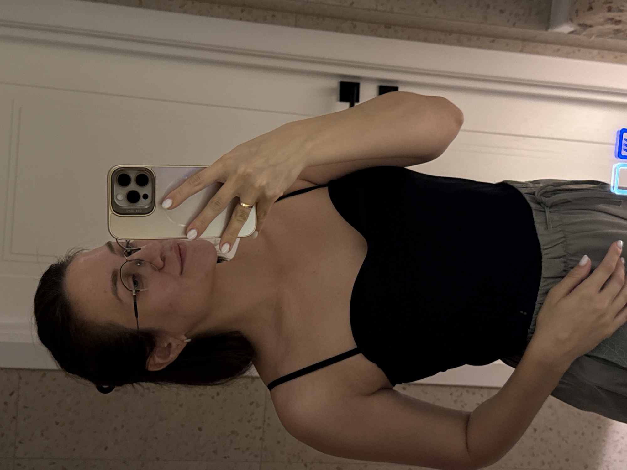 A young woman with dark hair pulled back and wearing eyeglasses is taking a selfie in a bathroom. She is wearing a black spaghetti strap top and grey pants. She is holding a smartphone in her right hand, and her left hand is resting on her lap. The bathroom appears to be modern, with tiled walls and a white door. There are no explicit signs of the city of Pervomaysky, Russia, in the image.