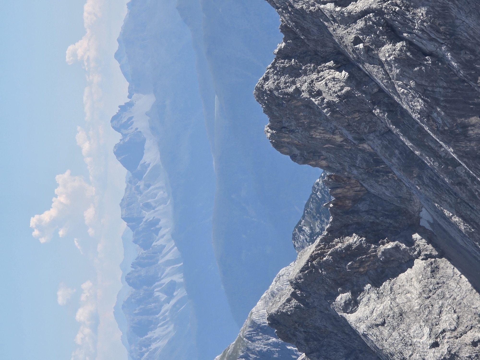 The image shows a layered view of mountains under a clear blue sky with scattered white clouds. The foreground features rugged, rocky peaks with sharp, angular formations and visible texture. In the mid-ground and background, successive ranges of mountains recede into the distance, becoming softer and more muted in color due to atmospheric perspective, appearing in shades of blue. The scene is a natural landscape, likely a mountainous region, given the topography. The location is identified as Garmisch-Partenkirchen, Germany. The image is a static landscape photograph with no people, animals, or discernable objects of human construction. The environment is characterized by natural geological formations and atmospheric conditions.
