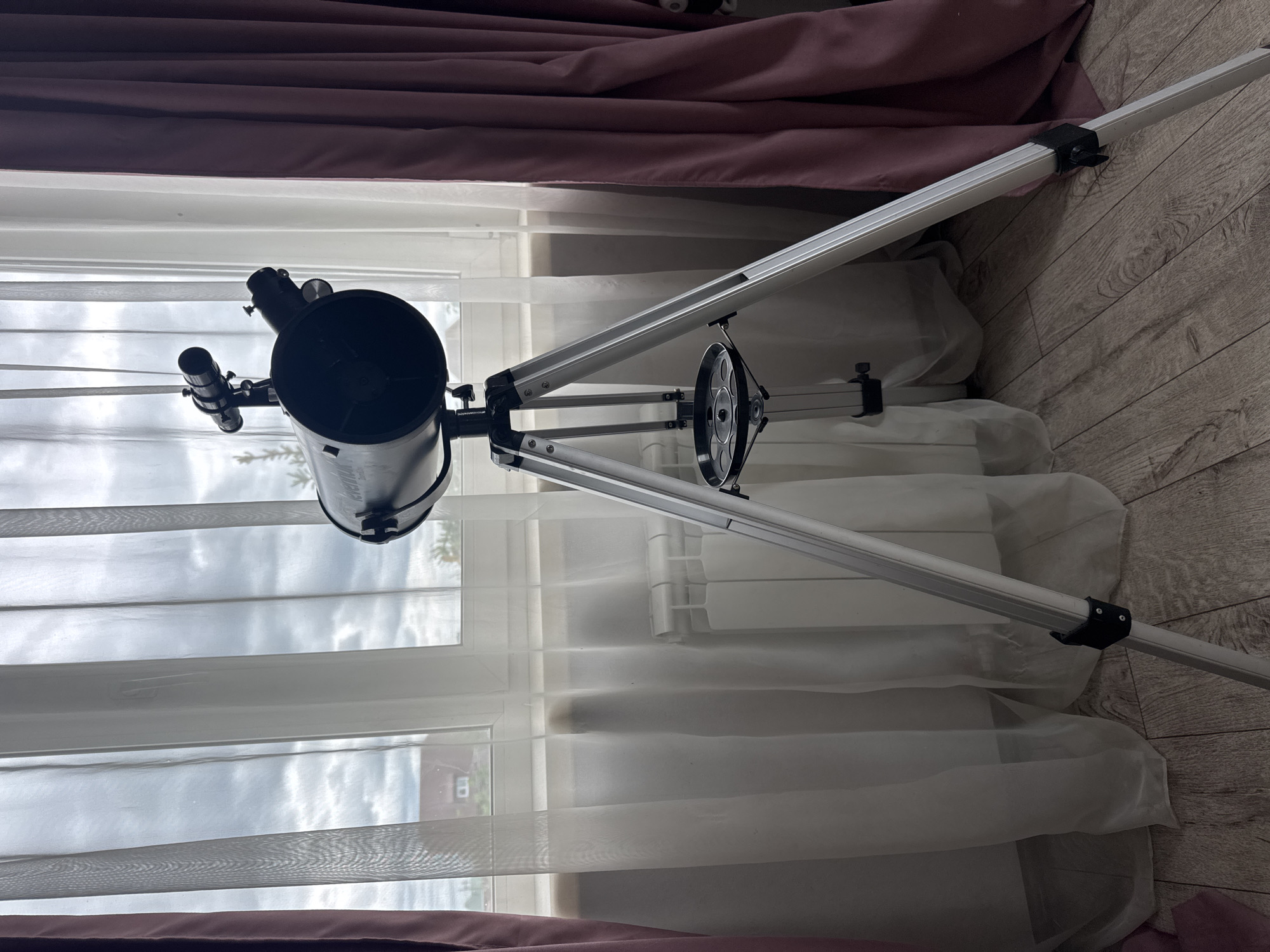 The image depicts a telescope set up on a tripod indoors, positioned in front of a window with sheer white curtains and darker pink curtains on the sides. The window overlooks a cloudy sky and a house with a red roof in the distance, suggesting an outdoor scene in Khomutovo, Russia. The telescope is black and silver, with a smaller finder scope attached to the main tube. A circular accessory tray is mounted on the tripod. The floor is made of light-colored wooden planks. A white radiator is visible behind the sheer curtains. There are no people or animals in the image.