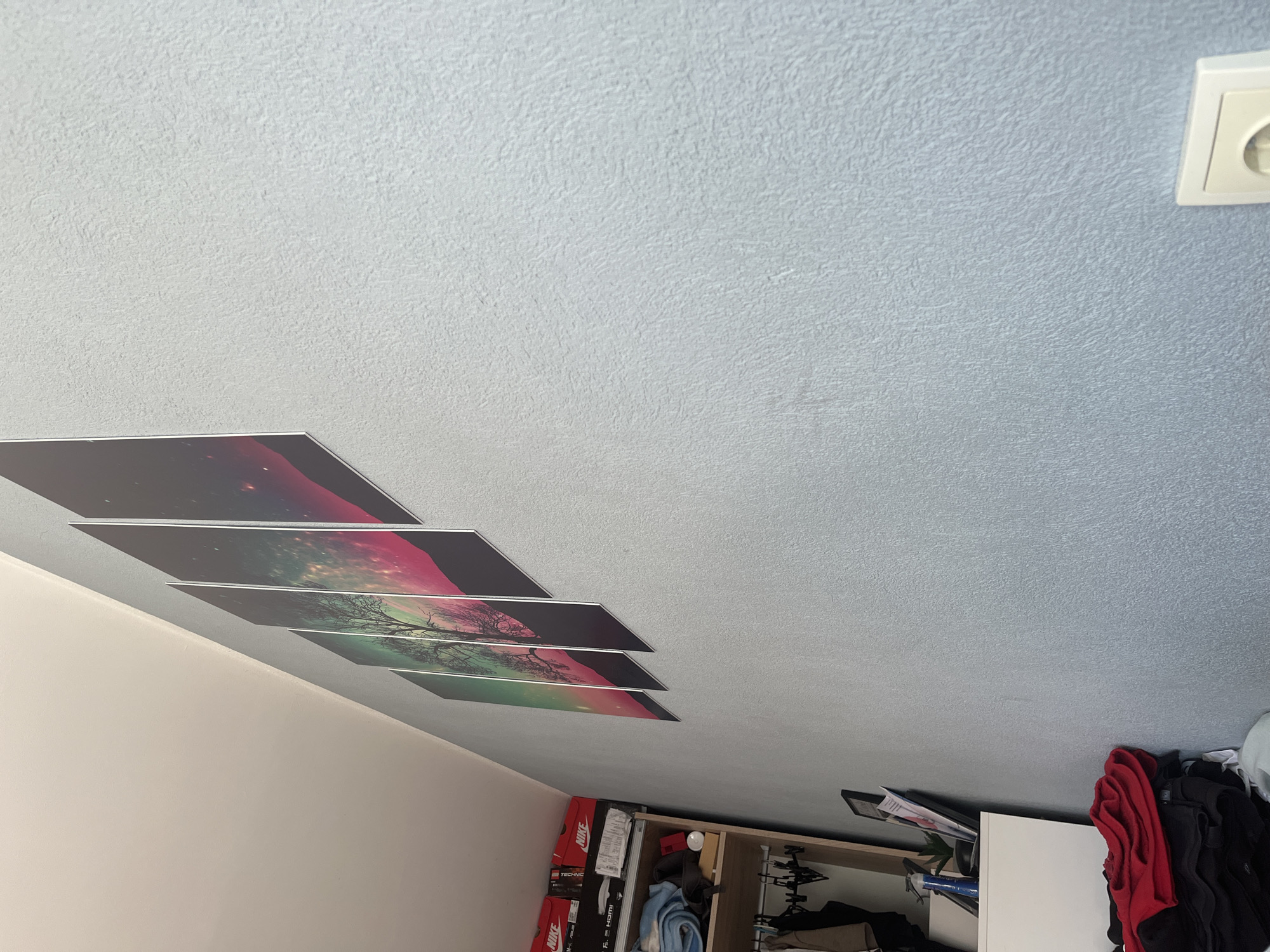 The image displays an interior room, likely a bedroom or living space, captured from a low angle looking upwards, creating a dynamic diagonal composition. The majority of the frame is occupied by a light blue-grey ceiling with a textured finish, resembling stucco or wallpaper. A smooth, light beige wall is visible along the bottom-left edge of the image.
Five rectangular art panels are mounted diagonally across the intersection of the ceiling and wall. These panels form a continuous panoramic image of a silhouetted tree with intricate branches, set against a vibrant, cosmic-themed gradient background. The background transitions from deep pink and purple tones to green and teal, with scattered points of light suggesting stars or distant nebulae. The panels are evenly spaced, creating a segmented effect for the artwork.
In the bottom-right corner of the image, household furniture and items are visible. This includes a white cabinet or dresser, and an open wardrobe or shelving unit. Inside and on top of these units, various clothes in red, black, and blue are neatly stored. Several shoe boxes are present, with visible branding including "Nike" and "TECHNIC," as well as "HDMI" on another box. Some papers with unreadable text are also stacked. A white electrical outlet is positioned on the textured ceiling in the top-right corner of the frame.
The lighting appears soft and even, possibly from an unseen window or artificial source, casting subtle shadows from the art panels onto the ceiling. There are no people visible, and no activity is taking place. The scene depicts a well-organized and decorated personal space, located in Pokrovnik, Bulgaria.