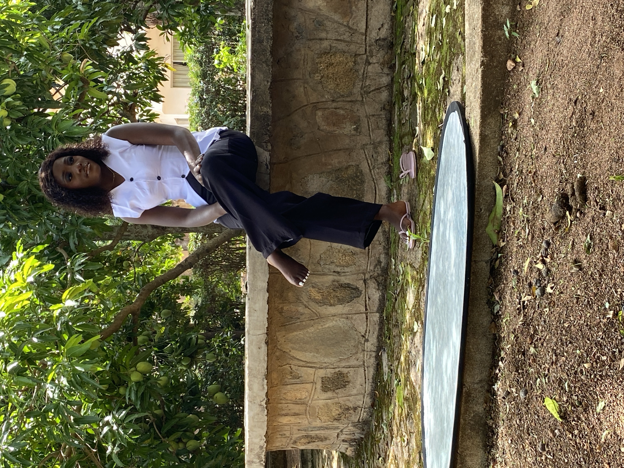 The image depicts a woman sitting on a stone ledge in an outdoor setting. She is wearing a white top and black pants. She appears to be barefoot with pink sandals next to her. A reflective board is placed on the ground next to the ledge. Behind the woman is a tree filled with green mangoes. The ground is covered with dirt and leaves. Based on the presence of mangoes and architectural details, the location is presumed to be Bukuru, Nigeria.
