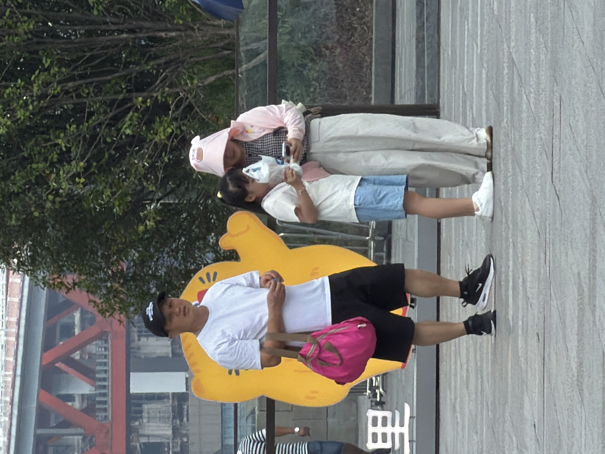 A man and a young girl stand outdoors near a yellow cartoon cat cutout in Yuzhong District, China. The man, wearing a white t-shirt, black shorts, and a black baseball cap, holds a bright pink bag with green straps. The girl, dressed in a white t-shirt and blue shorts, stands to the right of the man. Beside her, a woman in a pink hooded jacket and light-colored pants is looking at her phone. The background features a large red bridge structure and lush green trees.