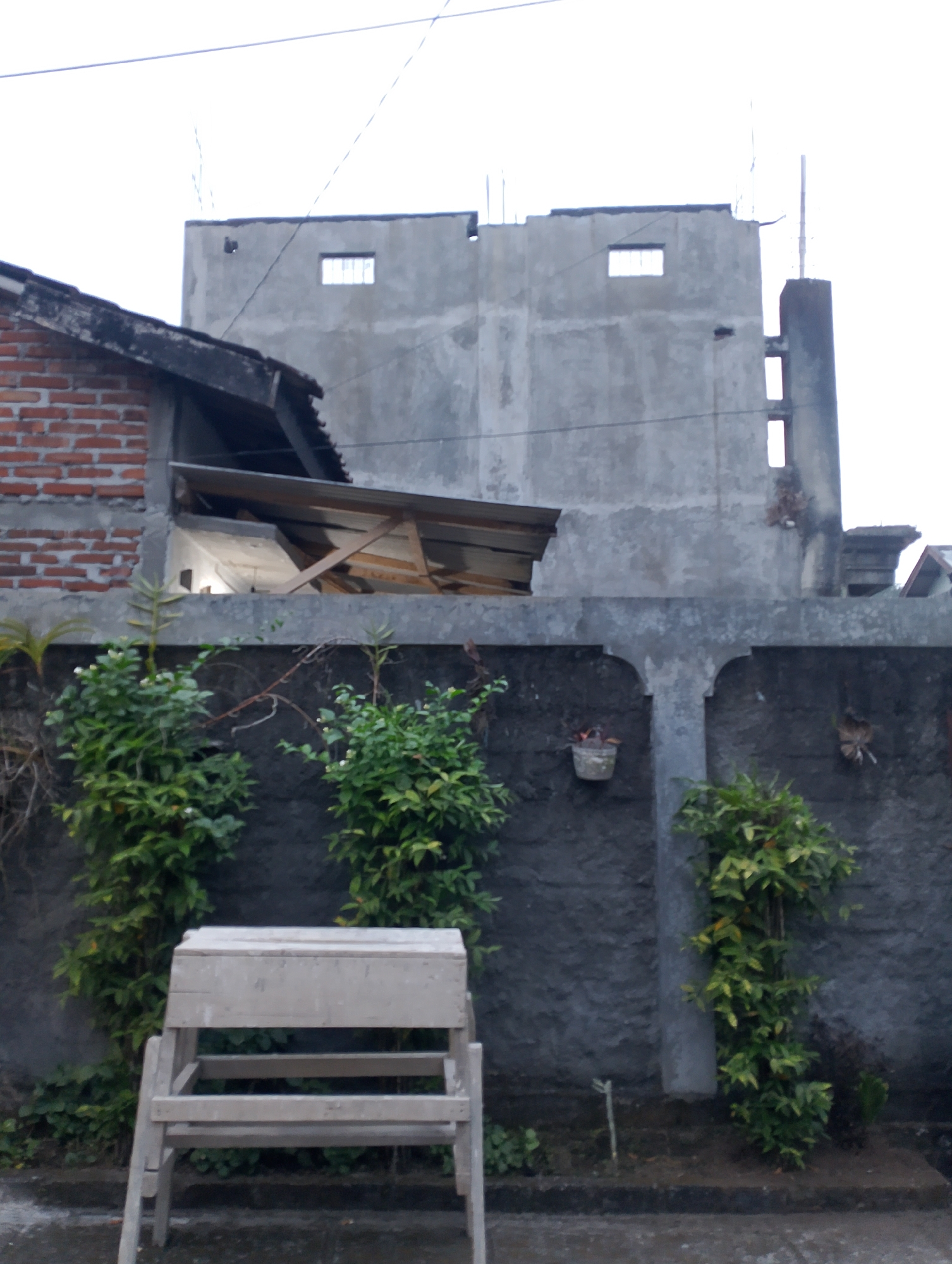 A low-angle, full outdoor shot captures a partially constructed building in Payaman, Indonesia, with a concrete wall in the foreground. A simple wooden bench sits in front of the wall. The building features exposed brickwork on the left, transitioning to unfinished concrete construction. Three rectangular windows are visible on the concrete facade, two in the upper portion and one elongated window lower down. Lush green plants climb the wall, some with small white flowers. A small hanging planter is attached to the wall on the left, and another hanging object, possibly a decorative item or a bird's nest, is on the right. The sky is overcast, suggesting a cloudy day.