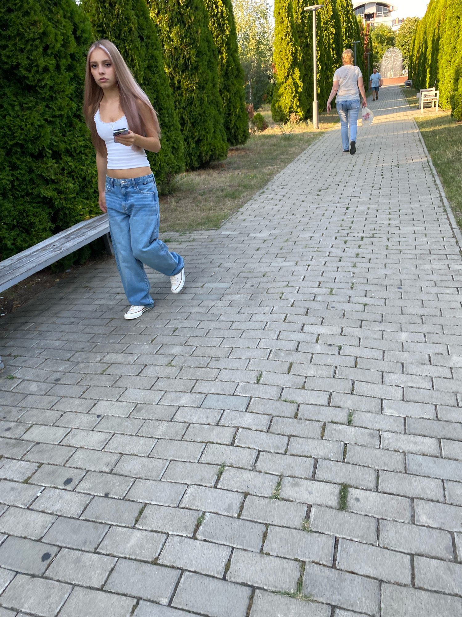 A young woman with long, blonde and pink streaked hair and a white tank top stands on a paved walkway, holding a smartphone. She is wearing loose-fitting blue jeans and white sneakers. A wooden bench is visible to her left. In the background, another woman in a light blue shirt and jeans walks away from the camera, carrying a plastic bag. The scene appears to be a park or public area with rows of tall green trees lining the walkway. The paving stones are light gray and rectangular. The overall lighting suggests it is daytime. The location is not specified in the image or crops.