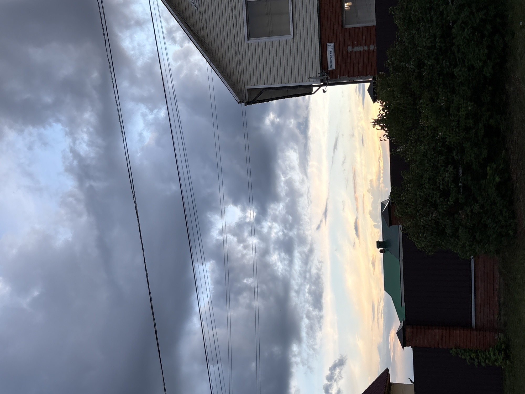 The image shows a view of a streetscape in Прикамский, Russia. The sky is filled with heavy, gray clouds, indicating an overcast day. Power lines stretch across the left side of the frame. On the right, the corner of a building with white siding and a window is visible, along with a brick building next to it. A street sign reads "Пионерская 12." The side of a tree or bush is visible next to the building.