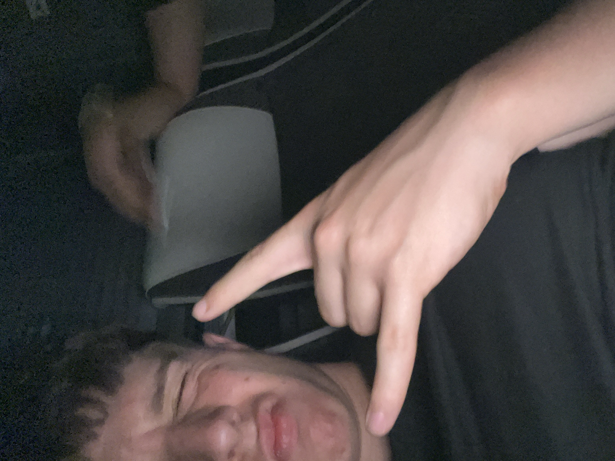 The image shows a person lying down, with another person's hand making a "devil horns" gesture near their face. The background is dark and indistinct, but appears to include clothing and possibly a chair. The faces of both individuals are partially obscured. It is not possible to determine if the location is Dinkata, Bulgaria, as there are no discernible landmarks or identifying features.