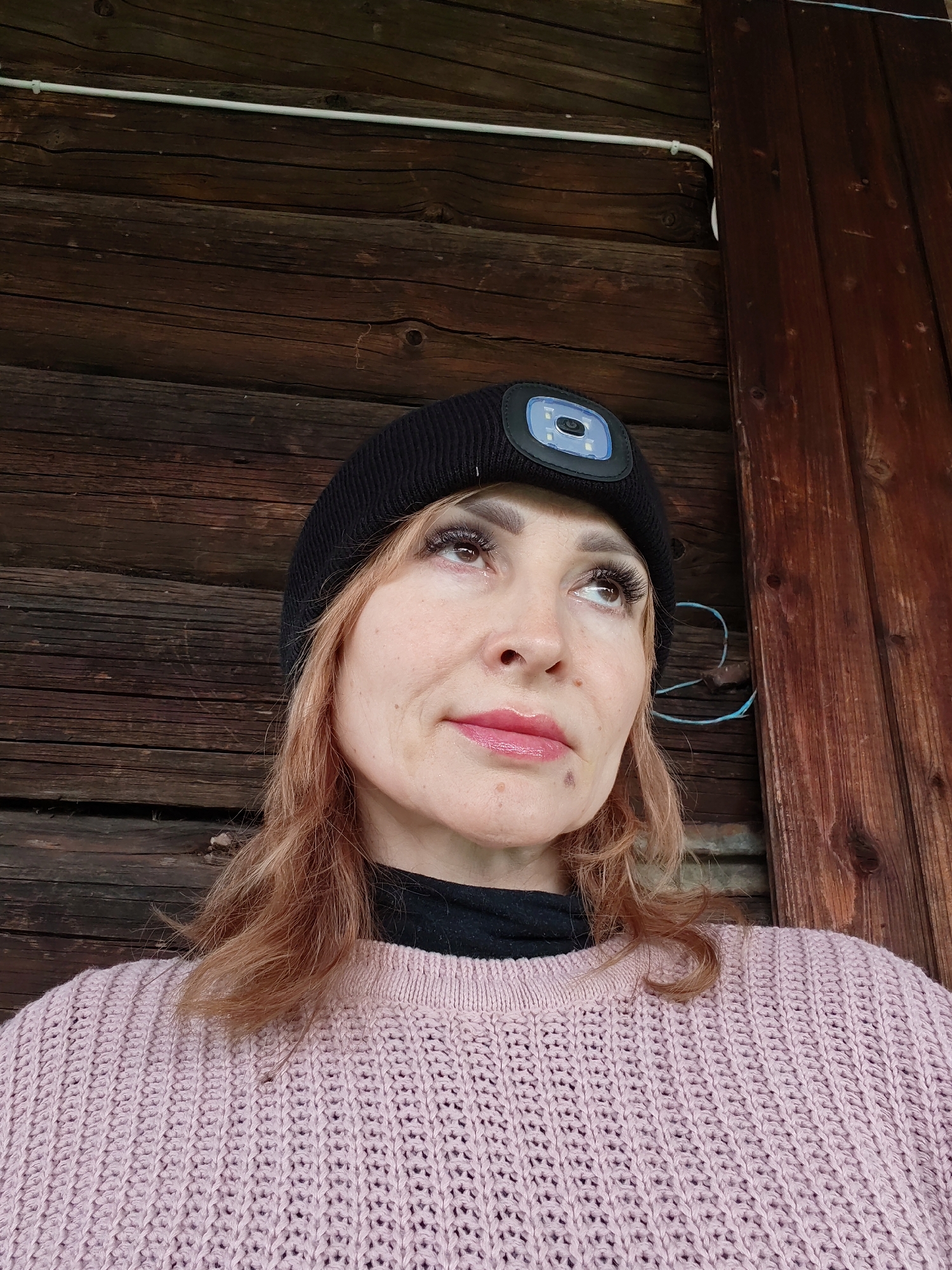 A woman stands in front of a wooden wall in the city of Chaikovsky, Russia. She is wearing a pink knitted sweater, a black turtleneck, and a black beanie with a headlamp attached. The wall behind her appears to be made of rough-hewn wooden planks. A white electrical conduit runs horizontally along the wall near the top of the frame. A blue wire is also visible attached to the wall. The woman has reddish-blonde hair and is looking up and to the left. She has artificial eyelashes and her lips appear to be glossy. A mole is visible on her left cheek.