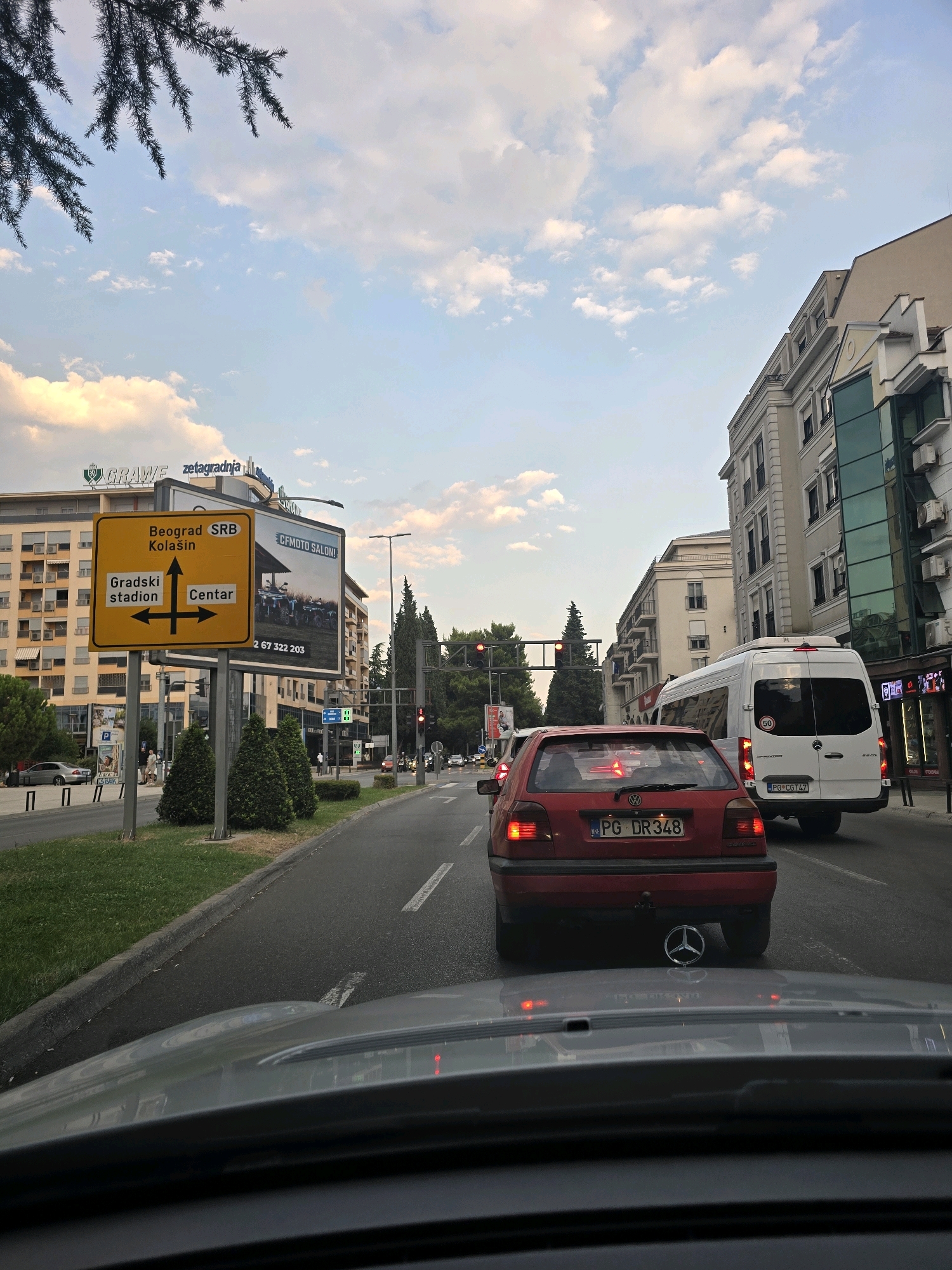 The image shows a street scene in Podgorica, Montenegro. In the foreground, the hood of a car is visible, suggesting the photo was taken from inside another vehicle. A red Volkswagen Golf is in front, with its license plate reading "PG DR348". The car has a Mercedes-Benz emblem on its exhaust pipe. To the right of the Golf, a white Mercedes-Benz Sprinter van is driving, with a license plate "PG CG747". A yellow traffic sign indicates directions to "Beograd SRB", "Kolašin", "Gradski stadion", and "Centar". Buildings line the street, with one prominent building on the left displaying the "GRAWE zetagradnja" logo. The sky is partly cloudy with a soft, diffused light. The overall impression is of a city street with moderate traffic, likely during the late afternoon or early evening.