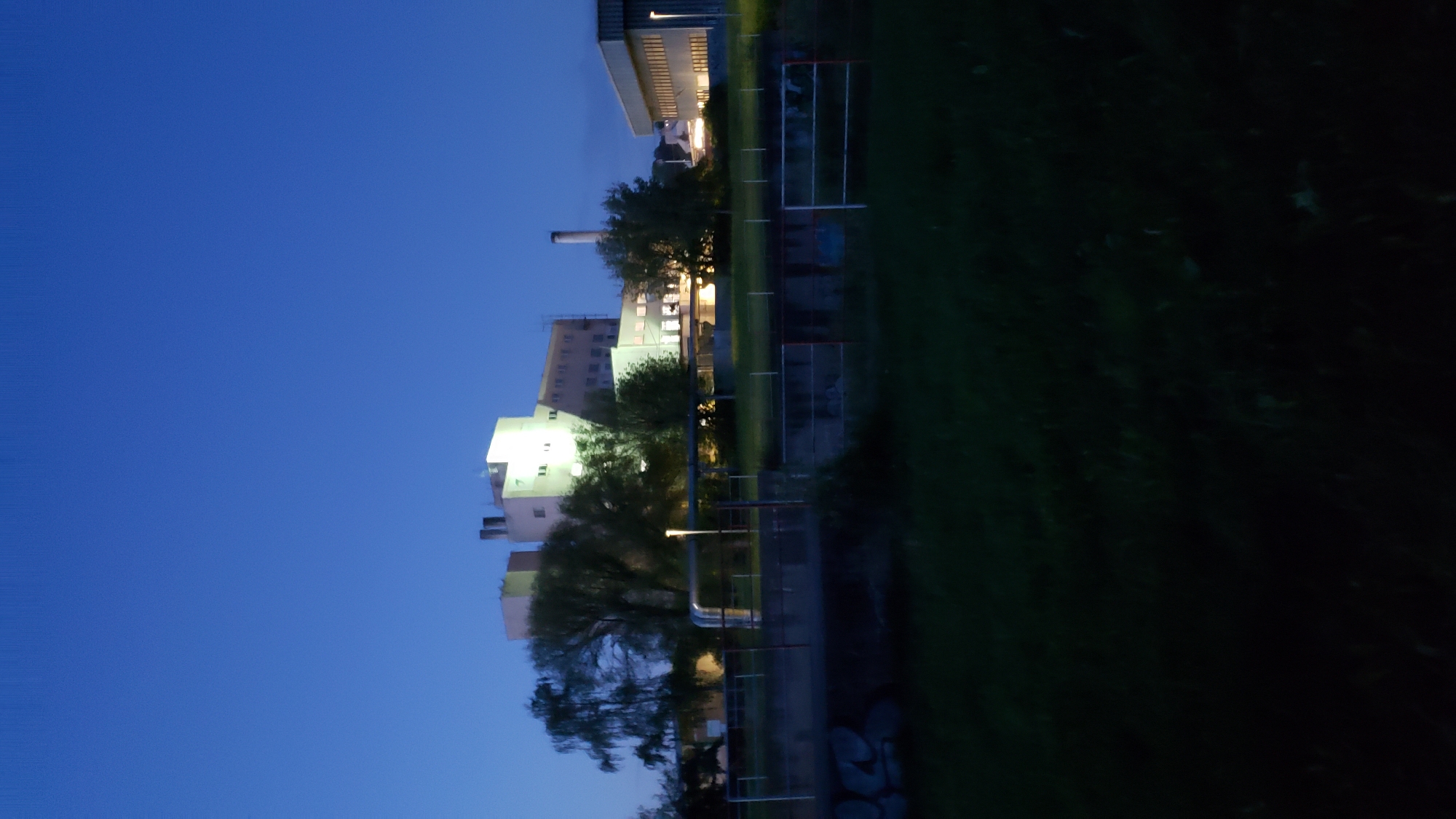 The image, taken at dusk, shows a modern, multi-story building complex with some of its windows illuminated. The architecture suggests an industrial or institutional use, possibly a factory or research facility, situated in Prešov, Slovakia. The building is surrounded by trees, some of which are bare, indicating it may be autumn or winter. In the foreground, a grassy area leads to a concrete structure with metal railings and fencing, suggesting a containment or boundary. The sky is a deep blue, characteristic of twilight. There are no people visible in the frame.