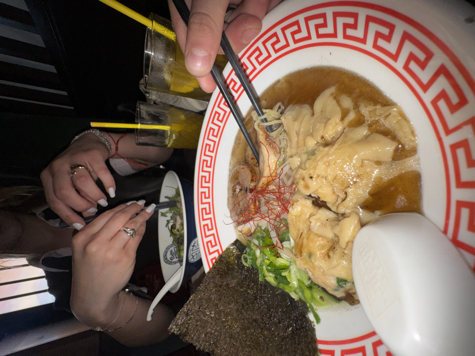 This image captures a close-up, high-angle view of a dining scene within an indoor restaurant, contextually located in Paris, France. The primary focus is on two bowls of food and the hands of two individuals engaging with their meals.
In the immediate foreground, a white bowl with a distinctive red Greek key pattern along its rim contains a large serving of ramen. The ramen broth is a rich brown, holding various ingredients including wavy noodles, several large, light-colored dumplings or wontons, slices of what appears to be pork belly (chashu), finely chopped green onions, and sparse red chili threads. A large, dark green, rectangular sheet of nori seaweed is positioned vertically on the left side of the bowl. A white ceramic ramen spoon rests on the left rim of this bowl. A right hand, belonging to an unseen individual, holds black chopsticks, actively lifting a strand of ramen noodle from the bowl.
To the upper left, partially visible from the chest upwards, is a female. Her hands, adorned with silver bracelets and rings on multiple fingers (including a prominent ring on her left ring finger), feature white nail polish. She is seen manipulating a fork and a white ceramic spoon within a smaller, blue and white patterned bowl containing green leafy ingredients, possibly a salad or a different type of soup.
Further to the right and slightly behind the main ramen bowl, two clear glasses filled with a yellow liquid and fitted with yellow straws are present on the table. The background is dark and out of focus, suggesting low ambient lighting typical of a restaurant setting. A soft, warm light source, potentially a window or decorative lighting, is visible behind the female. The scene depicts a shared meal between at least two individuals.