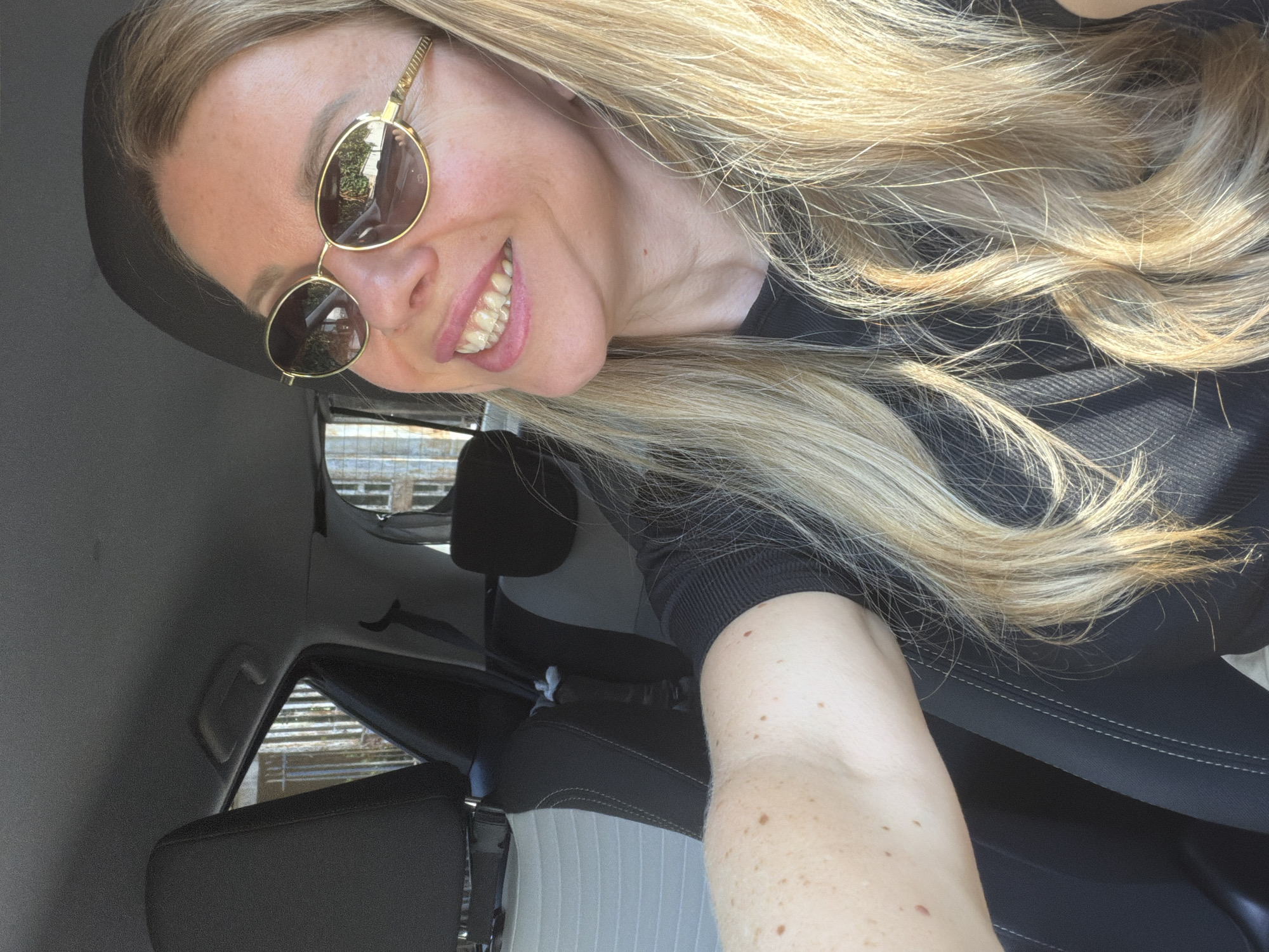 A blonde woman wearing sunglasses and a black top is taking a selfie in the passenger seat of a car. She is smiling broadly, revealing her teeth. The interior of the car, including the headrests and seatbelt, is visible. The woman's arm and shoulder are in the foreground, showing freckles. The background is blurred, but appears to be a street scene with some buildings and trees. The lighting suggests it is daytime, possibly in a sunny climate. There is no discernible information about the location being Rome, Italy.