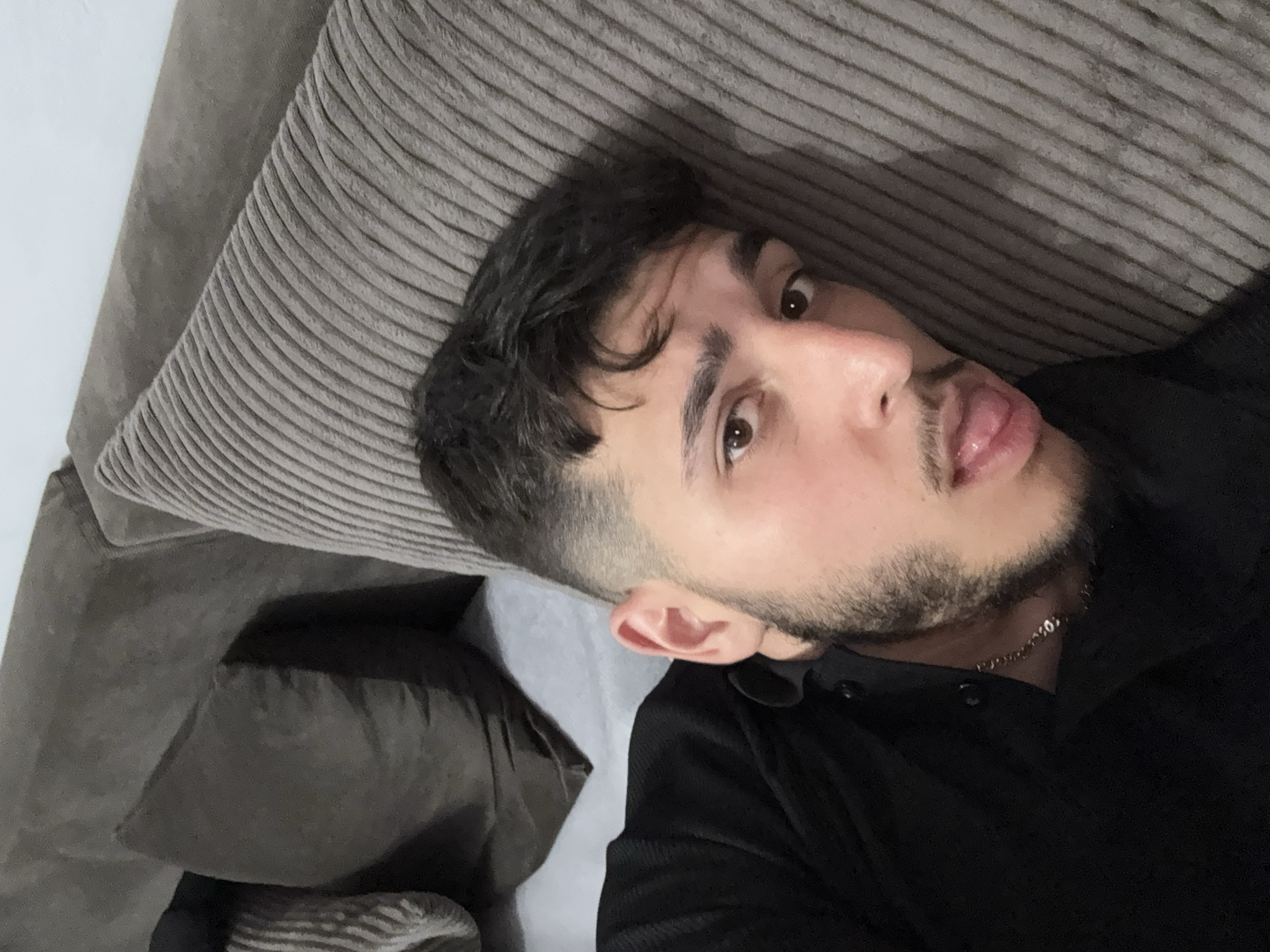 The image displays a young adult male lying on a sofa in an indoor environment, situated in the city of Varna, Bulgaria. The individual possesses dark, wavy hair with faded sides, dark brown eyes, a trimmed beard, and a mustache. He is wearing a dark, likely black, long-sleeved collared shirt, and a thin chain necklace is visible around his neck. His head rests on a light grey cushion with a distinct ribbed texture, similar to corduroy. To the left of the frame, a plain dark grey cushion is present, contributing to the sofa's appearance. The sofa itself is dark-colored. A light-colored, plain wall forms the background. The male is looking directly at the camera with his tongue slightly extended.