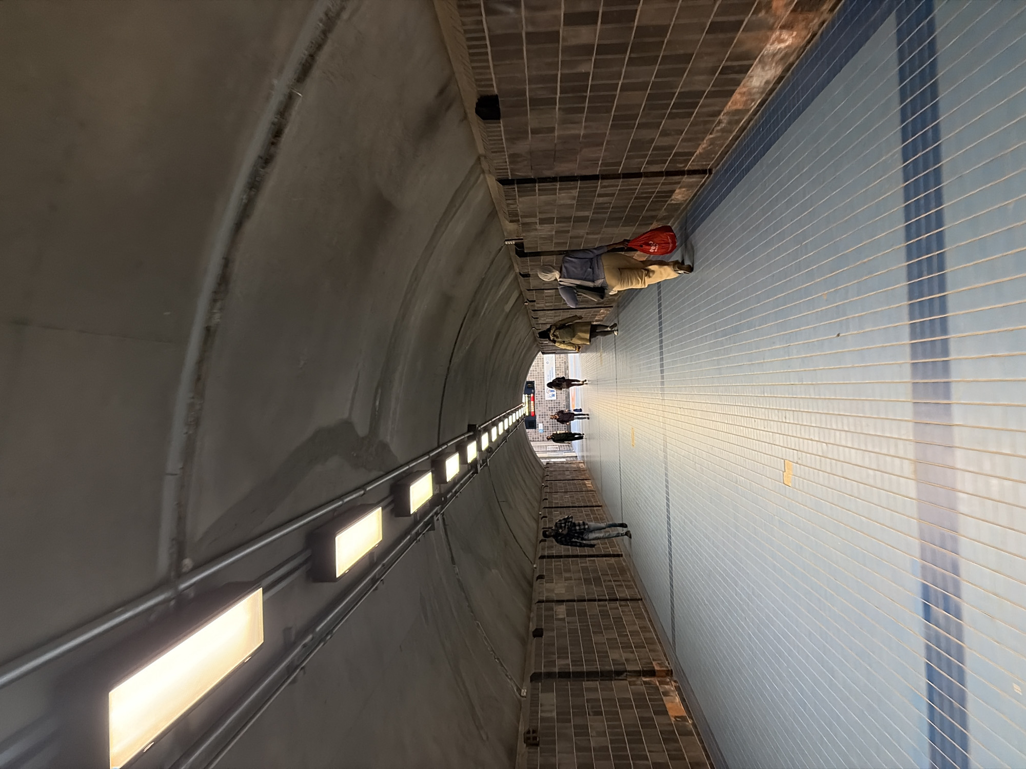 This is an indoor pedestrian tunnel in Montreal, Canada. The tunnel has an arched ceiling on the left side, with a series of evenly spaced rectangular lights running along the top of the wall. The right side of the tunnel consists of a tiled wall. The floor has a brick pattern. Several people are walking through the tunnel, appearing small and distant towards the tunnel's exit. One individual closer to the foreground is wearing a red bag and light-colored clothing.