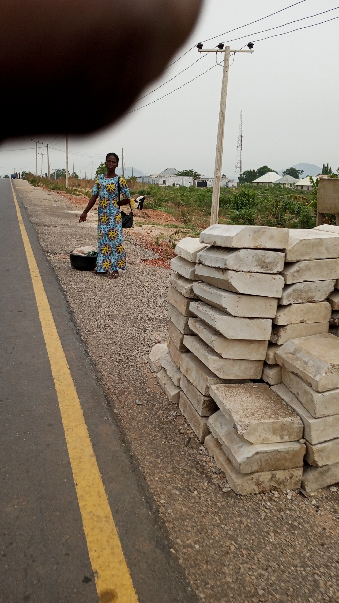 The image depicts a scene alongside a road in Adankolo, Nigeria. An African woman in a patterned blue and yellow dress stands beside the road, a black bag over her shoulder and a black plastic basin containing what appears to be laundry at her feet. To her right, adjacent to the road's edge, is a stack of rectangular, light-colored concrete pavers. The road itself is paved and marked with a yellow line. In the background are sparse buildings, trees, a cell tower, and what appears to be a mountain range in the far distance under an overcast sky. An electrical pole with power lines is visible behind the woman. Part of a person's hand obscures the view at the top of the frame.