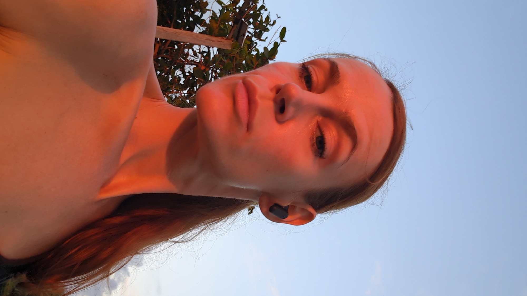 The media file is a close-up, rotated portrait of a young woman. She is outdoors, with a clear blue sky in the background and foliage visible to the left. The lighting suggests it is either sunrise or sunset, casting a warm, orange glow on her skin. She is looking up and slightly to her left, with her eyes open. She has on a black wireless earbud in her right ear. Her hair is long and reddish-brown, falling loosely around her shoulders. The image is framed in a way that emphasizes her face and upper body. There is no visible text in the media file. The setting is likely a natural environment, possibly a garden or park, given the presence of plants. The mood appears relaxed and contemplative. The location context is Mengwi, Indonesia.