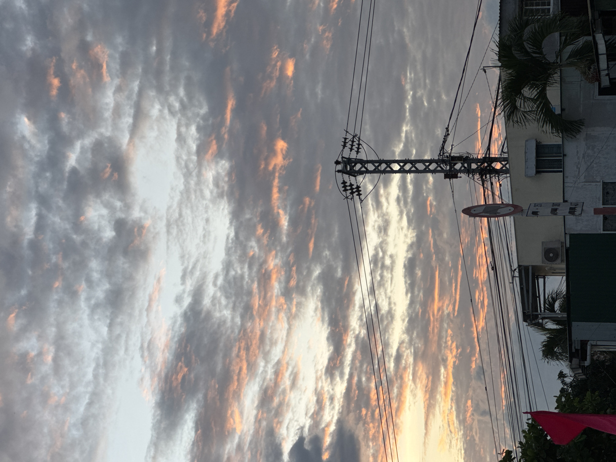 A picturesque sunset paints the sky over Nha Trang, Vietnam, with a dramatic display of clouds. The heavens are a canvas of gray, streaked with vibrant hues of orange and pink, suggesting the sun is either rising or setting. A prominent electrical tower stands tall, adorned with a complex network of wires that crisscross the sky. Below, a two-story building with a light-colored facade is visible, featuring windows and an air conditioning unit. To the left, a bright red flag flutters, partially obscuring some foliage. A traffic sign with a circular symbol and white lettering on a dark background is mounted on a pole. A palm tree adds a tropical touch to the scene. The overall impression is one of urban life bathed in the soft, ethereal light of dusk or dawn.