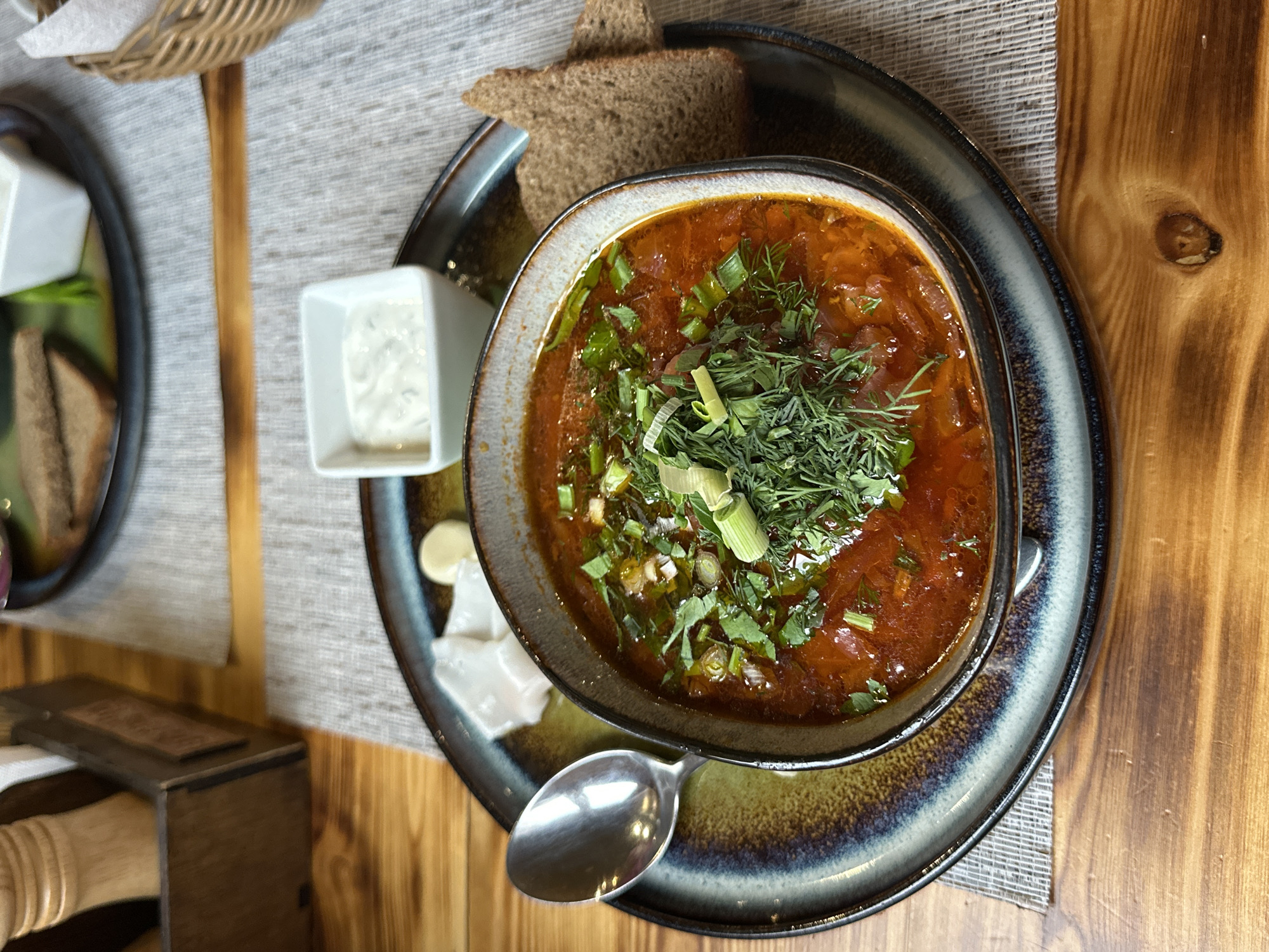 The image shows a bowl of soup, likely borscht, served on a dark-colored plate with a spoon. The soup is garnished with chopped greens and dill. Accompanying the soup are a small square dish of white sauce, pieces of bread, and what appears to be sliced cheese. The setting appears to be a restaurant, with a woven basket visible in the background and a wooden table. The location is Unknown, Unknown.