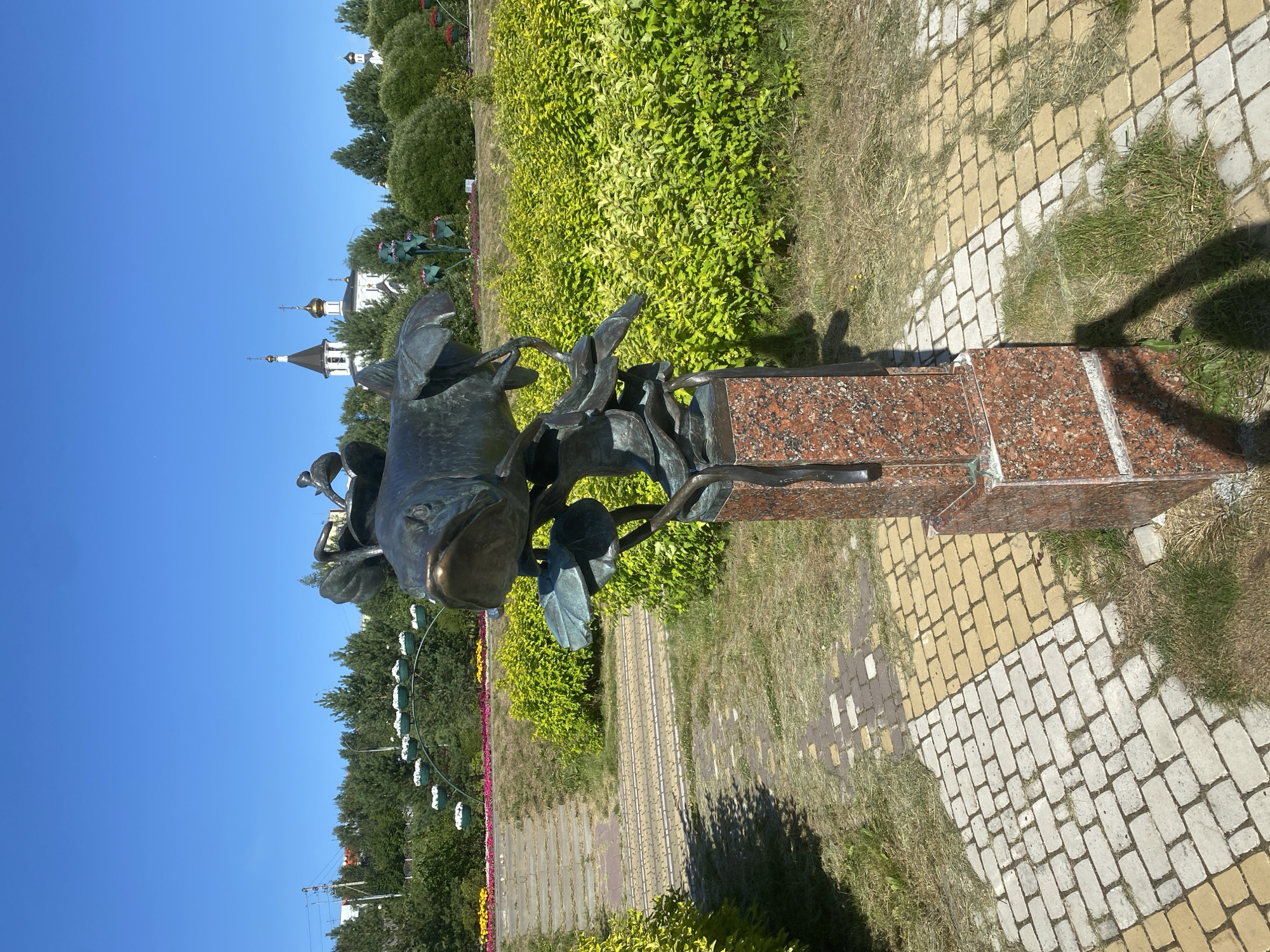 In Nefteyugansk, Russia, a bronze sculpture of a fish is prominently displayed in a park. The sculpture depicts a fish with large eyes, resting on what appears to be a lotus leaf, and is mounted on a red granite pedestal. The surrounding environment is characterized by lush green bushes and trees, with a clear blue sky overhead. In the background, several church domes are visible, suggesting the park's proximity to a religious complex. The pathways in the park are paved with light-colored and yellow bricks, creating a visually appealing mosaic pattern. A Ferris wheel-like structure with flower pots is also visible in the distance, adding to the park's decorative elements. The scene captures a bright, sunny day, with shadows cast by the sculpture and vegetation.