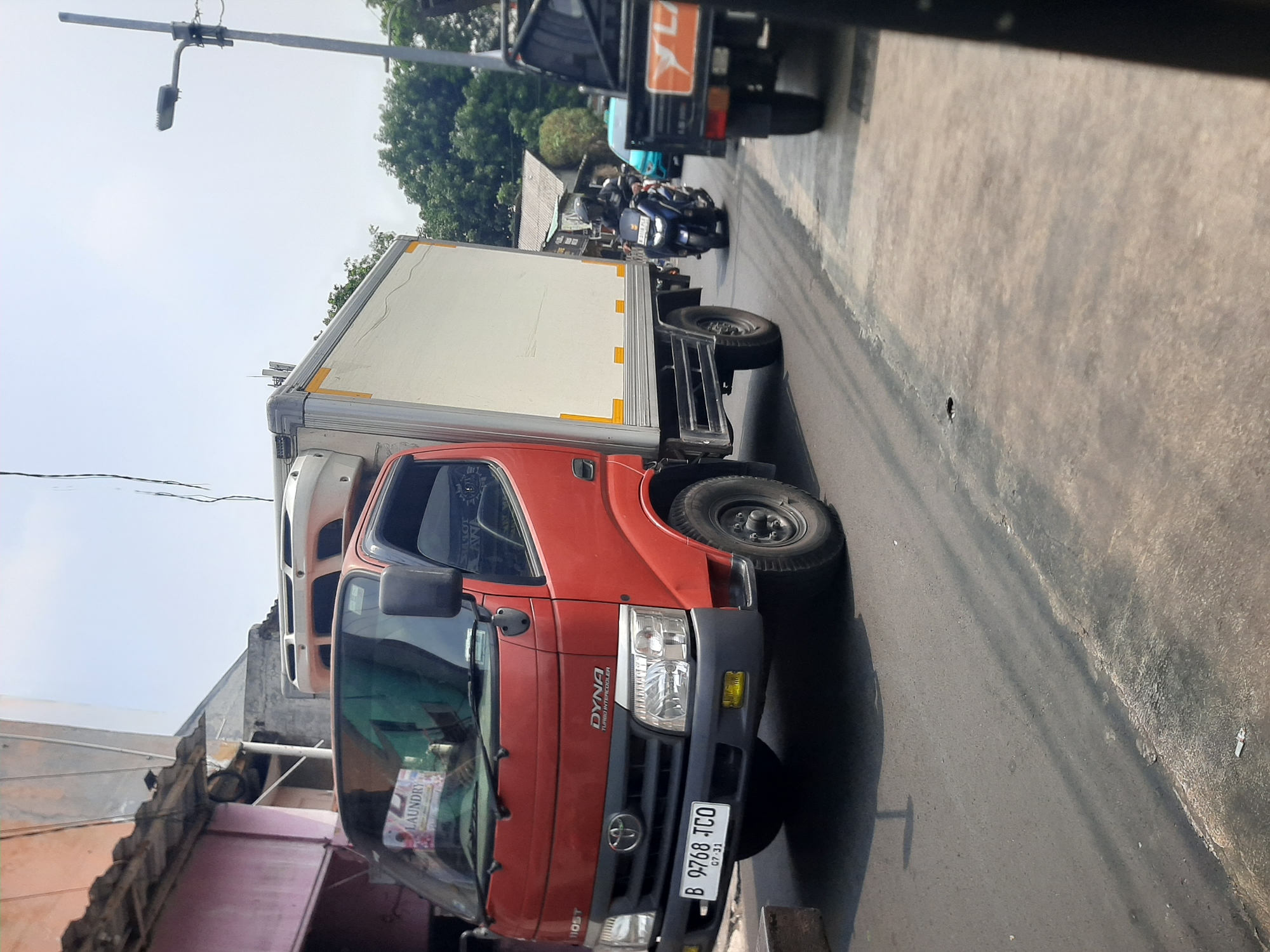 A red and white refrigerated truck, identified as a "DYNA TURBO INTERCOOLER," is parked on a street in Central Jakarta, Indonesia, with its license plate reading "B 9768 TCO 07-31." The truck is positioned on the right side of the road, adjacent to a building with a light pink facade and a corrugated metal roof. Behind the truck, a blue motorcycle with a rider wearing a helmet is in motion, followed by a teal-colored car. Further down the road, the rear of a black truck is visible. The environment appears to be an urban setting with overhead power lines and a street lamp. The day is bright with a clear sky and a few scattered trees in the background.