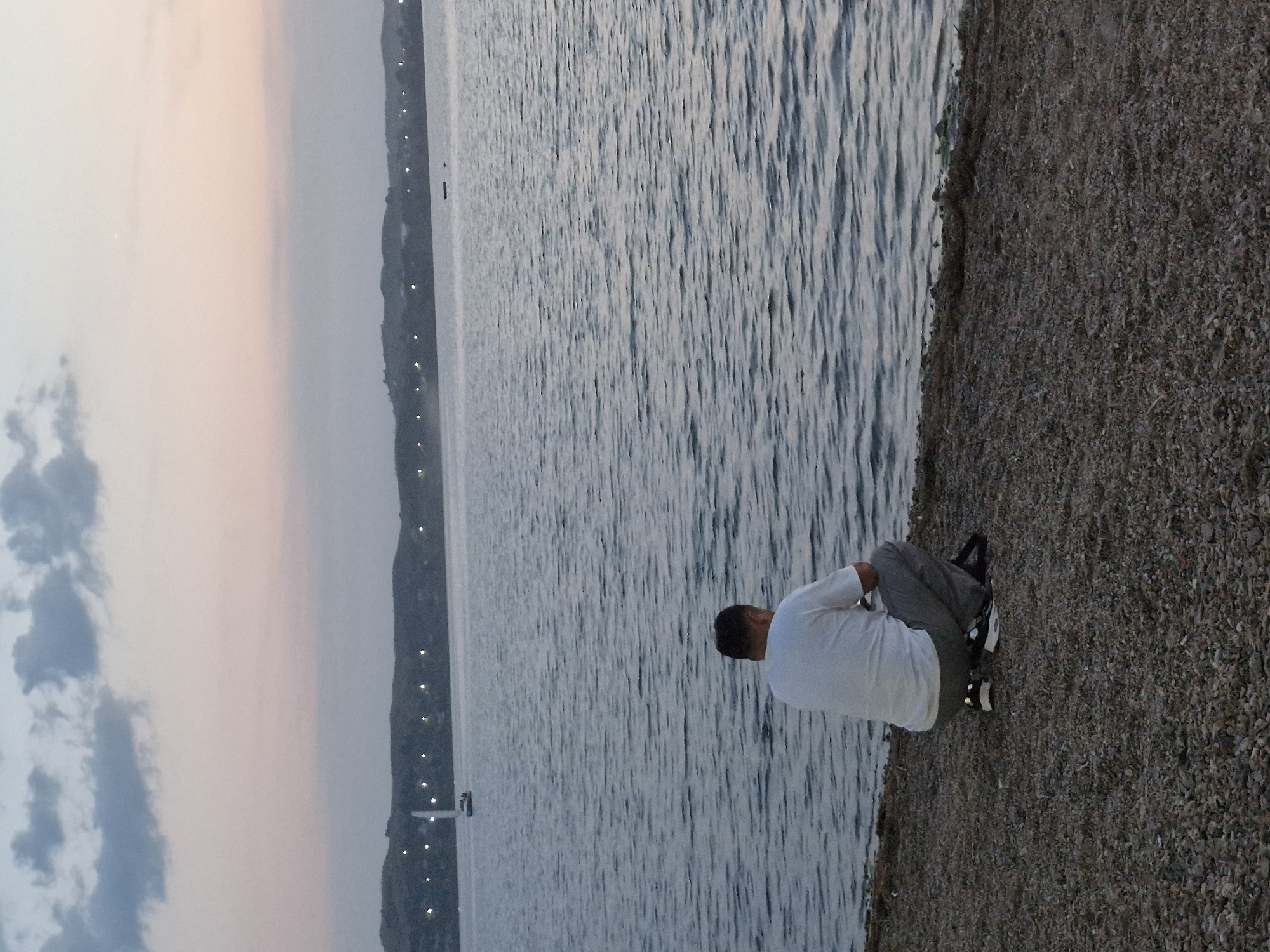 A person is sitting on the shore of a body of water, facing away from the camera. The person is wearing a white long-sleeved shirt and dark pants, with white shoes. The scene appears to be a lake or sea, with gentle waves. The shore is covered in small pebbles. In the distance, a silhouetted shoreline with lights is visible, suggesting a settlement or town. The sky is a soft gradient of pink and grey, indicating either sunrise or sunset. The overall mood is tranquil and contemplative. The image appears to be taken in Kokshetau, Kazakhstan. There is no visible text in the image.