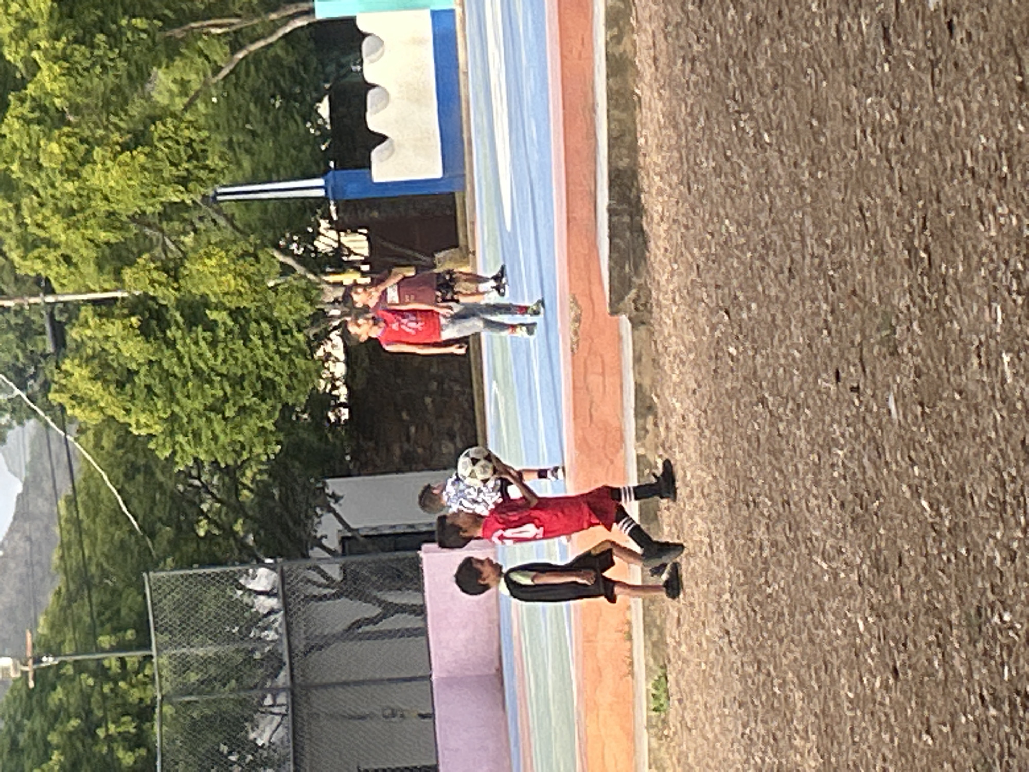 The image depicts an outdoor recreational area, likely a sports court, with a dirt/gravel foreground. In the mid-ground, a group of children are visible. The child on the left is wearing a black vest over a light-colored shirt and black shorts. The child next to him, in the center foreground, is wearing a red jersey with white stripes and black shorts, and is carrying a soccer ball. Another child in a patterned shirt is walking behind them. Further in the background, two more children, one in a red tank top and grey pants, and another in a dark shirt and patterned shorts, are standing on the court. The court itself has colorful markings, suggesting it is used for various sports. Behind the court, there are trees, a chain-link fence, and a building with a light-colored facade. In the far background, hills and mountains are visible under a bright sky. The overall setting suggests a warm, sunny day. This scene is consistent with a public park or school grounds in Santa Ana, Venezuela, where such sports facilities are common.
